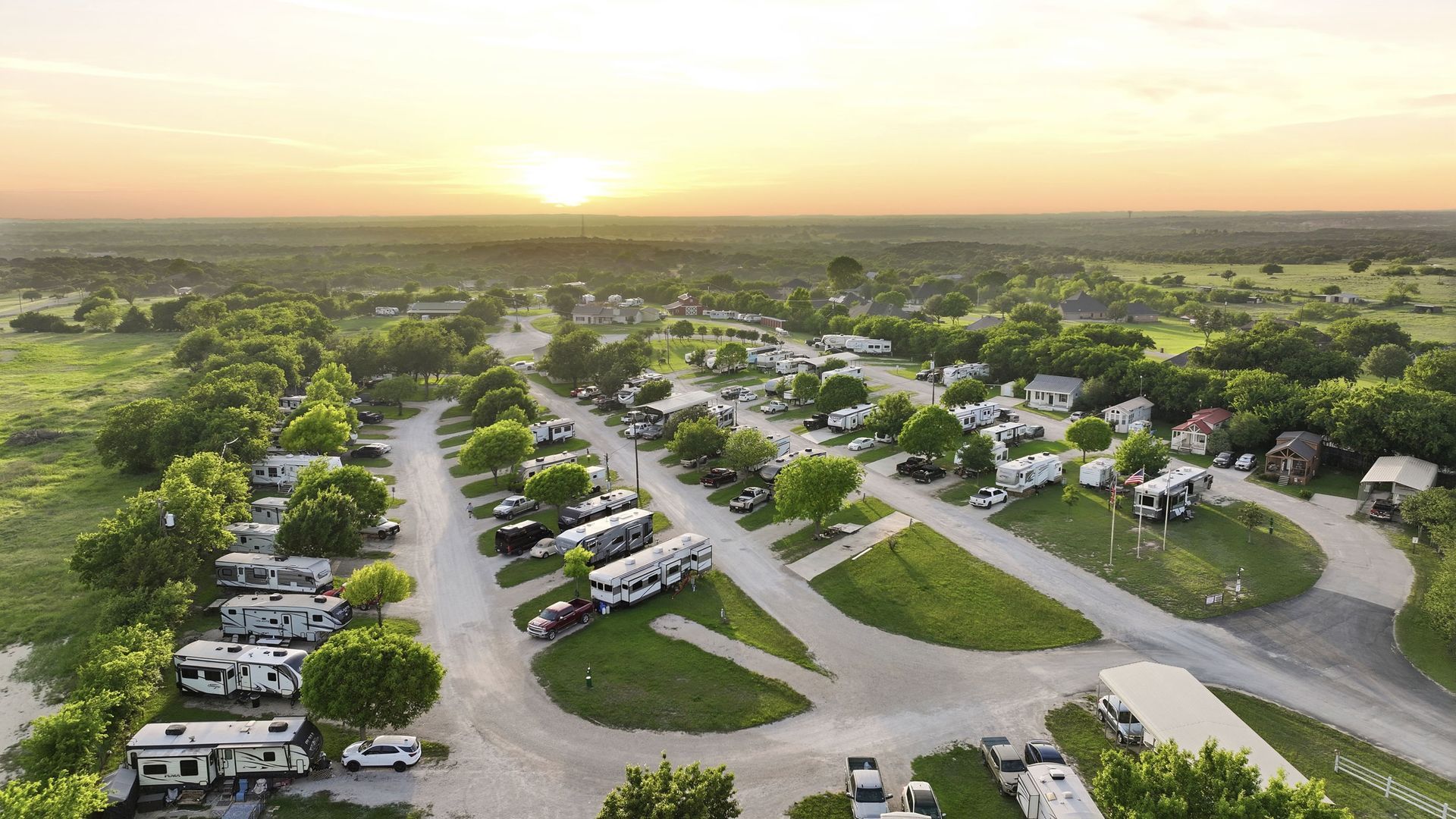 Aerial view of a suburban neighborhood at sunset with parked cars, roads, and green trees.