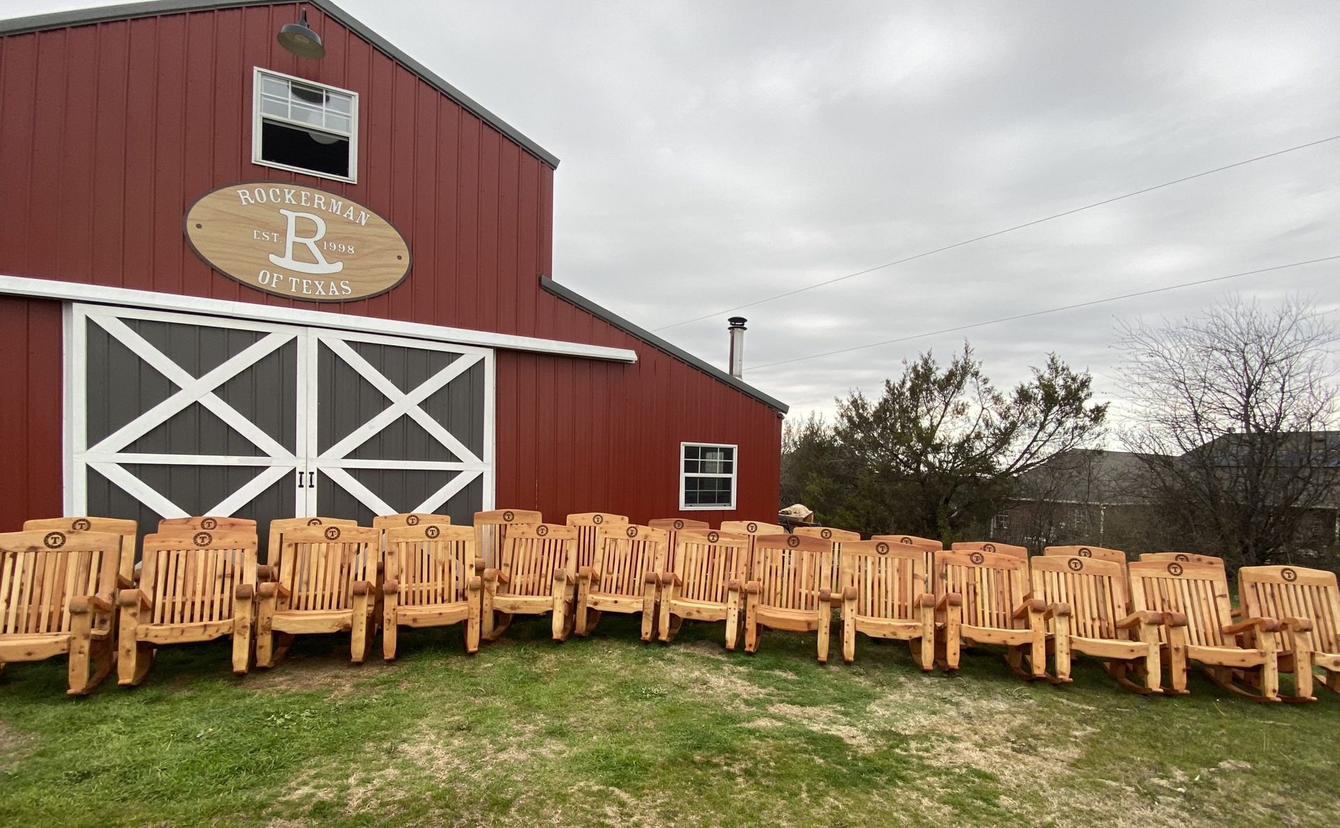 Red barn with white doors, lined with many wooden chairs, cloudy sky.
