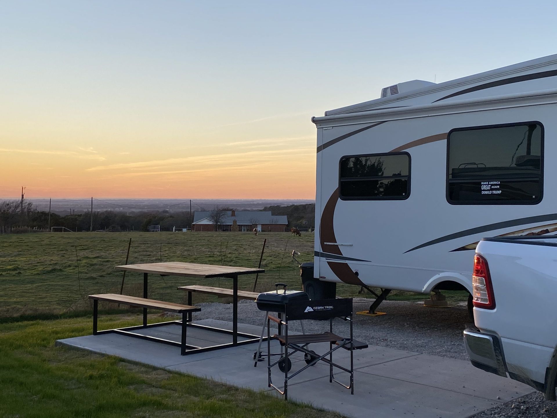 RV parked with picnic table and grill on a concrete pad, overlooking a scenic landscape at sunset.