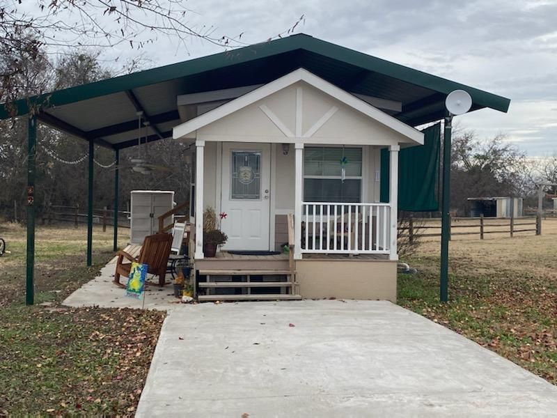 Small white house with porch under green carport. Concrete driveway, cloudy sky, rural setting.
