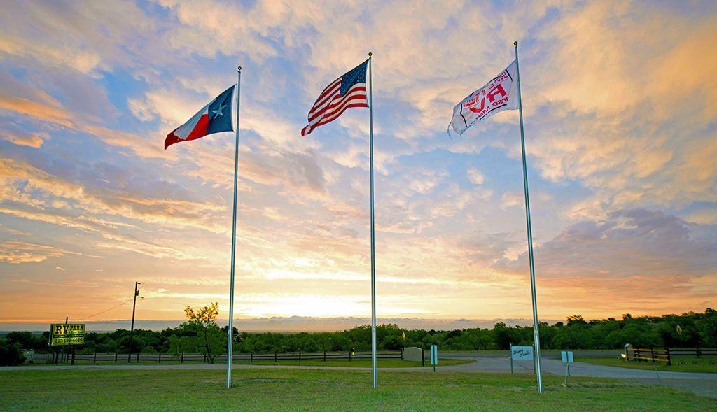 Three flags: Texas, USA, and a pink logo, fly in a field at sunset.