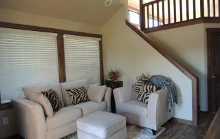 Living room with couch, chair, and ottoman. Beige walls, wood trim, blinds, and a staircase.