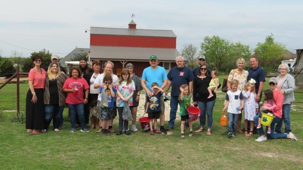 Large group of people pose in front of a red barn and fence on a grassy field. Several children hold Easter baskets.