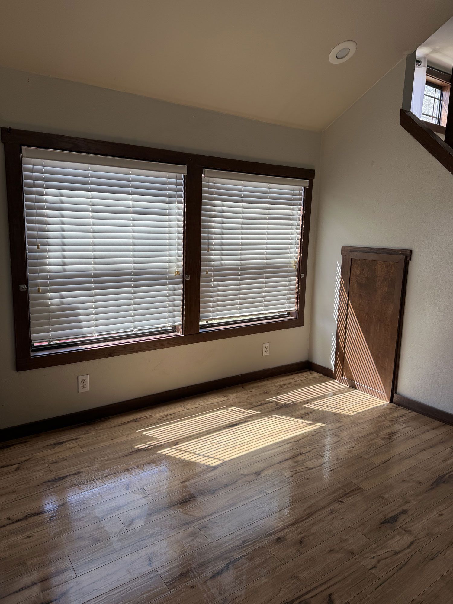 Empty sunlit room with hardwood floors, large windows with blinds, and a small door on the right.