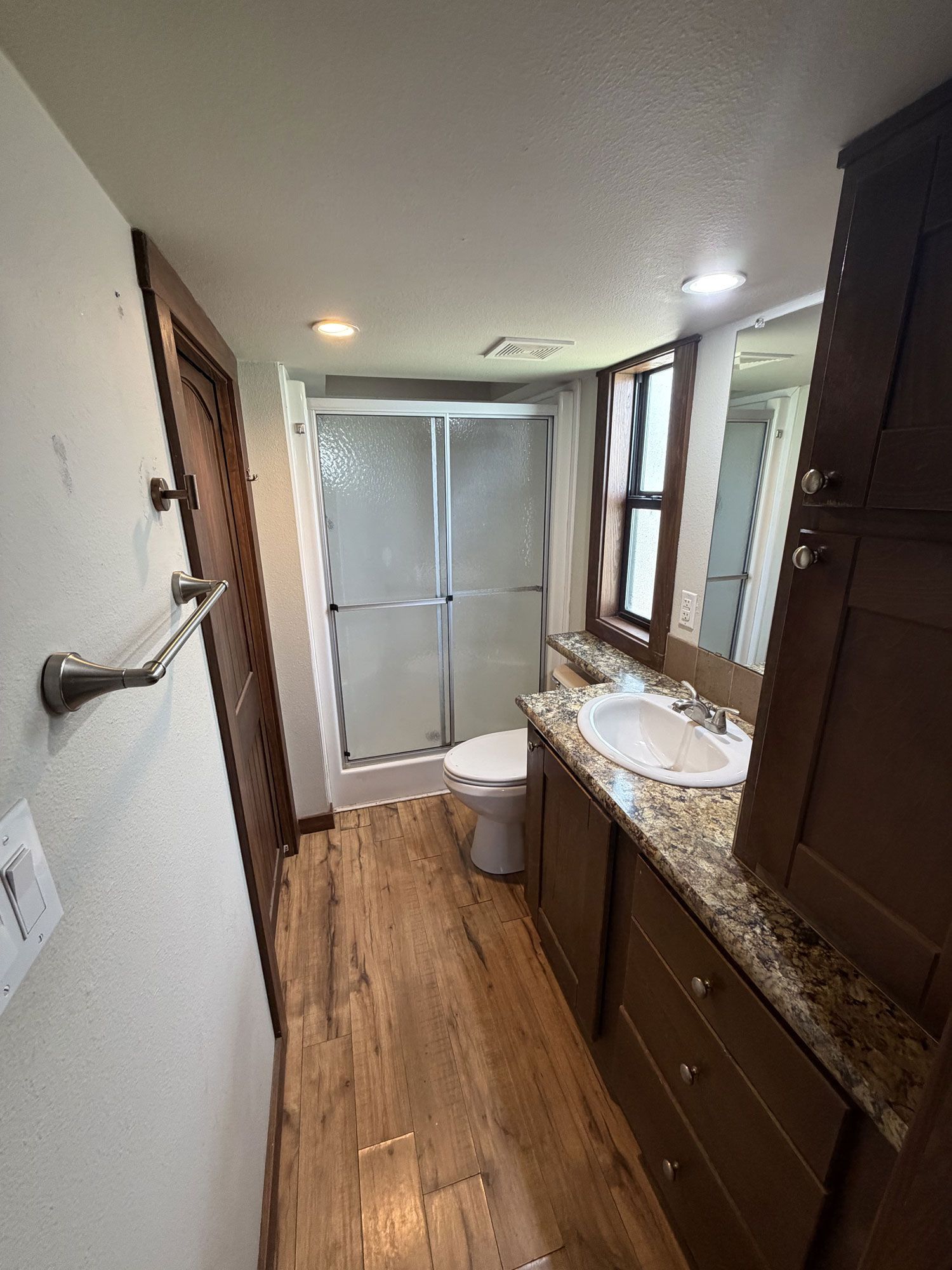 Narrow bathroom with wood vanity, granite counter, toilet, and frosted shower at the far end.