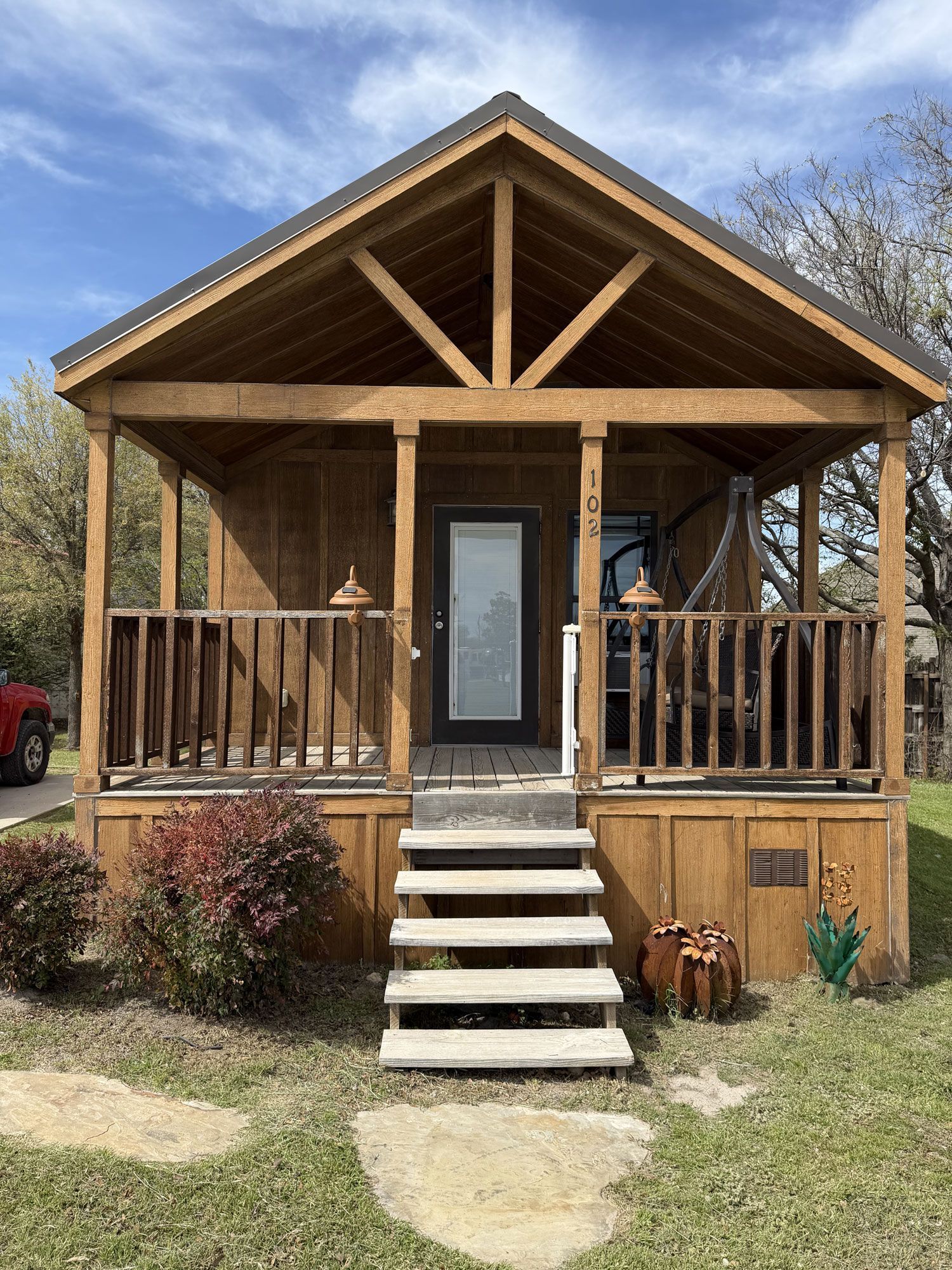 Small wooden cabin with a front porch, steps, railing, and swing, set in a grassy yard.