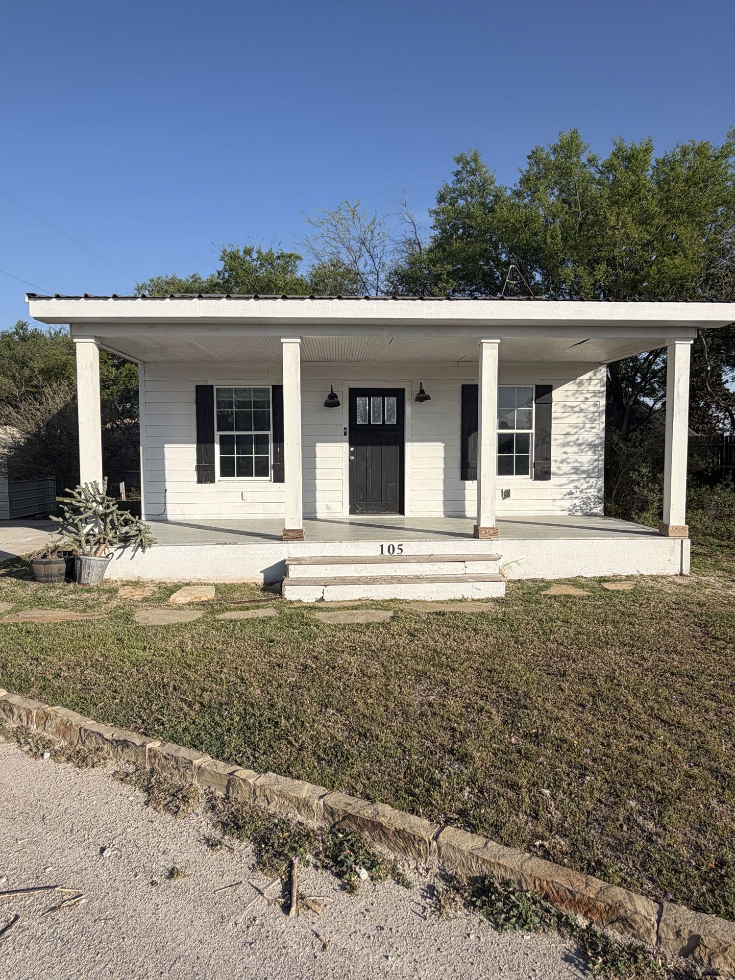 Small white house with a front porch and steps, set behind a grassy yard under a clear blue sky