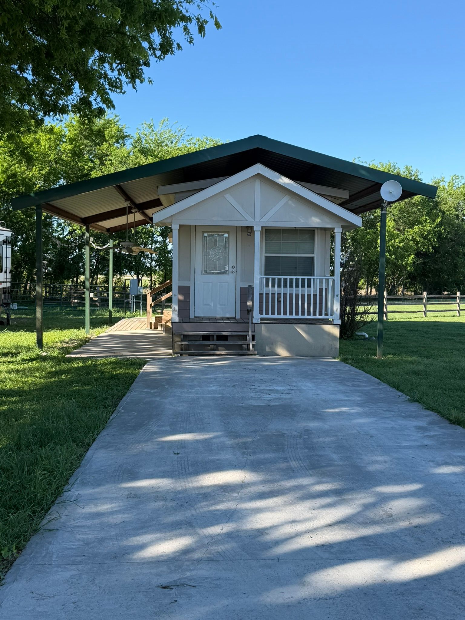 Small white cottage with a porch and long concrete driveway under trees in a sunny park-like setting