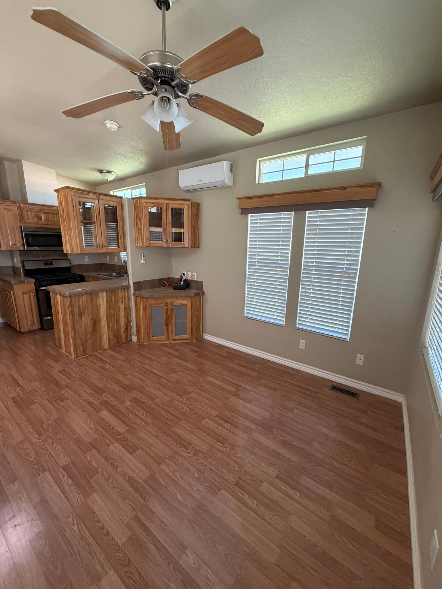 Empty living room with wood floors, ceiling fan, kitchen cabinets, and sunlit windows with blinds