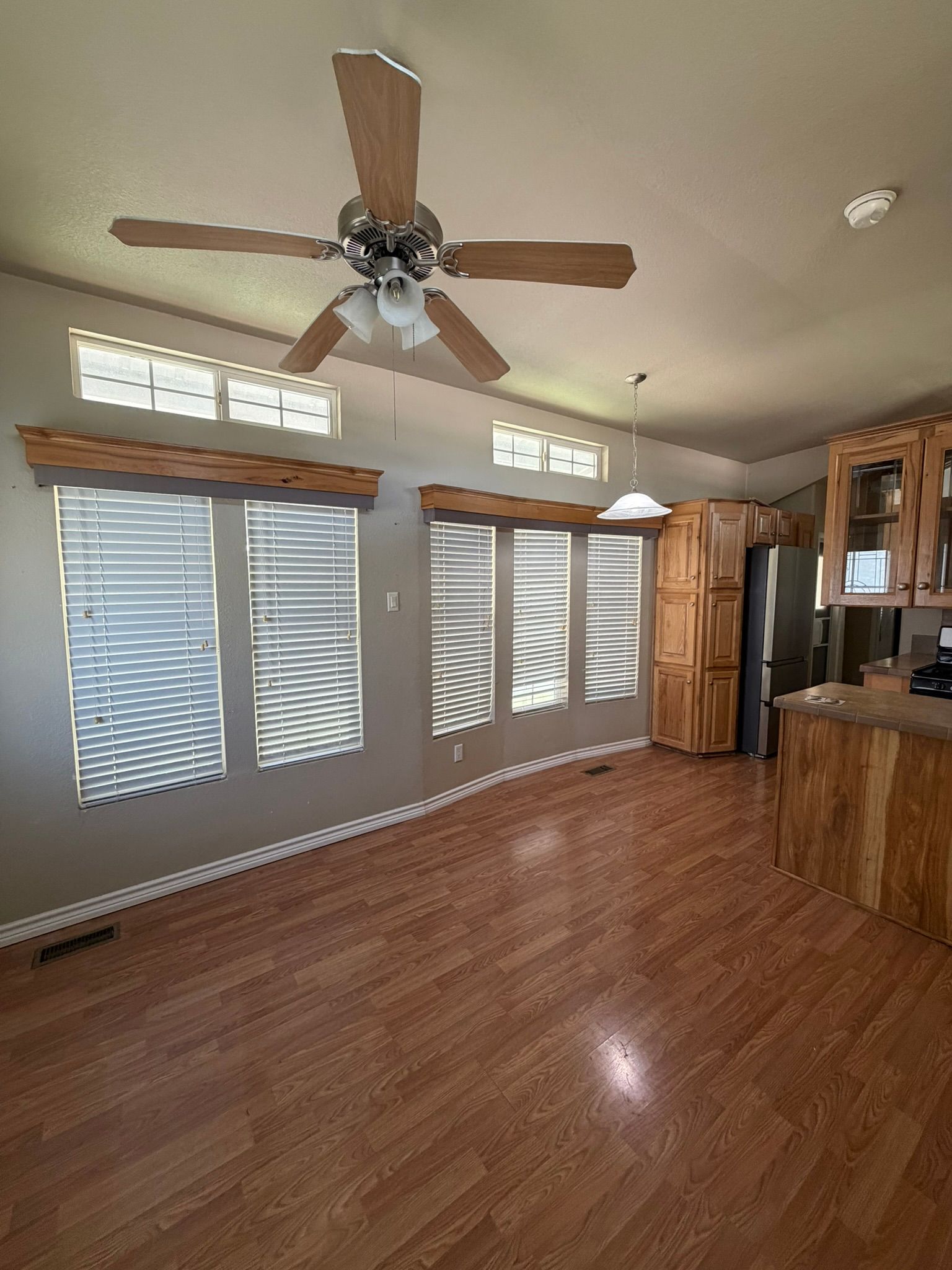 Spacious living room with wood floors, ceiling fan, large windows, and kitchen cabinets on the right.