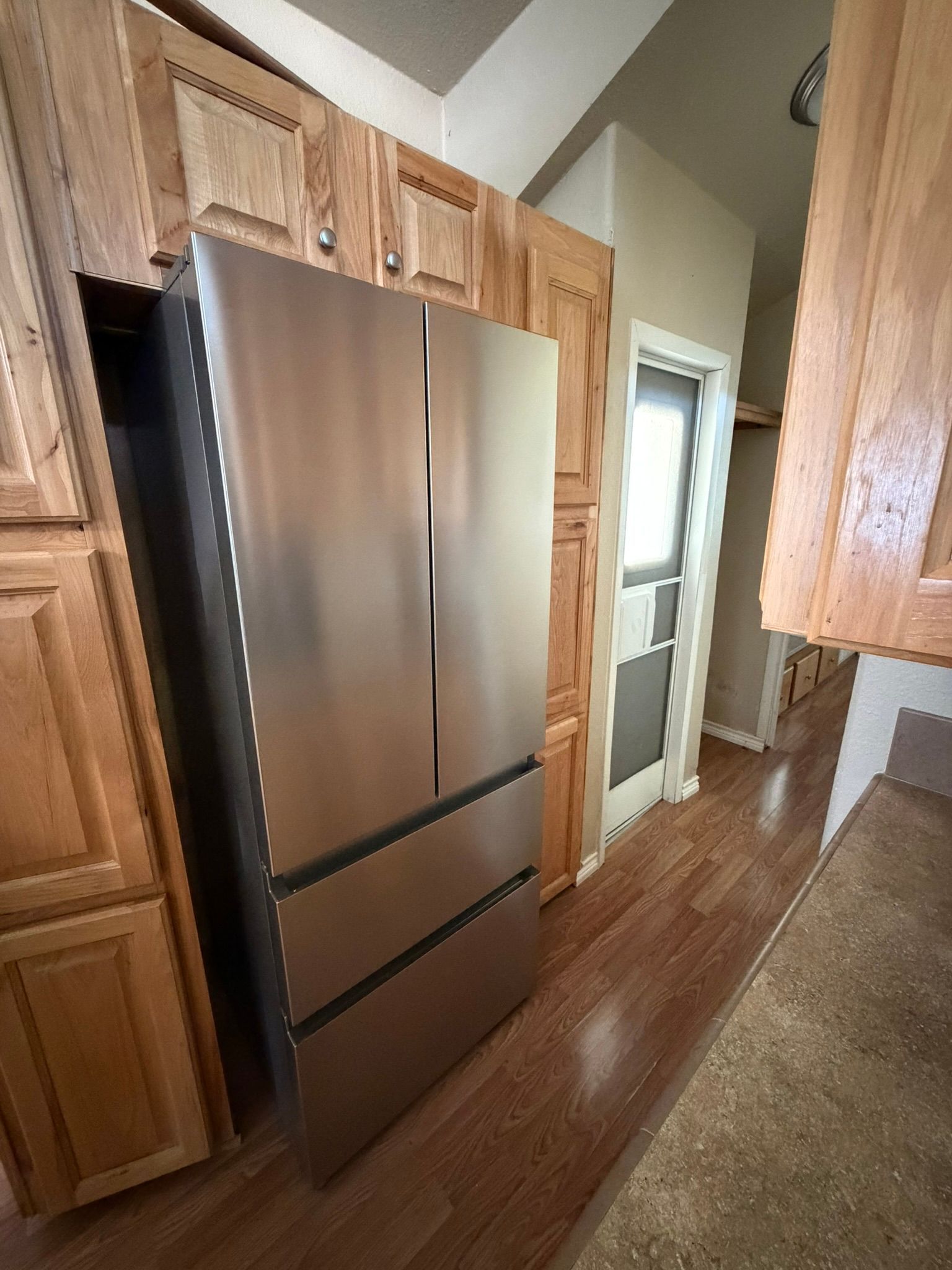 Stainless steel French-door refrigerator in a wood kitchen with cabinets and a hallway doorway nearby
