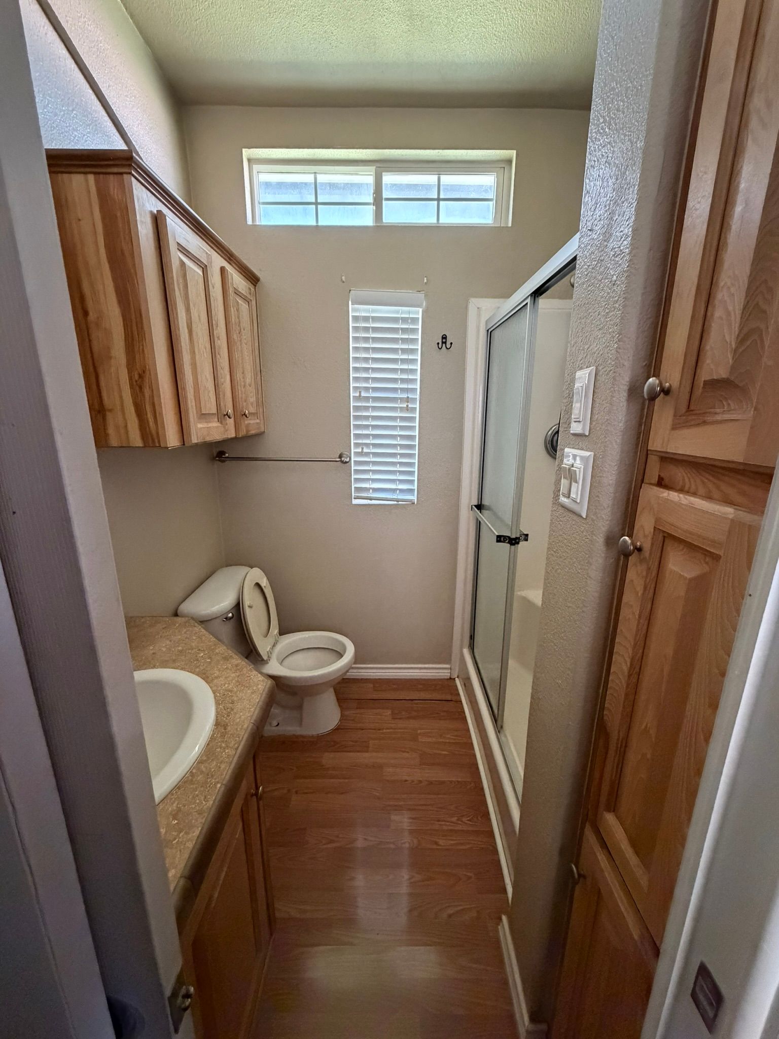 Narrow bathroom with toilet, sink, shower, and wood cabinets in a small hallway layout