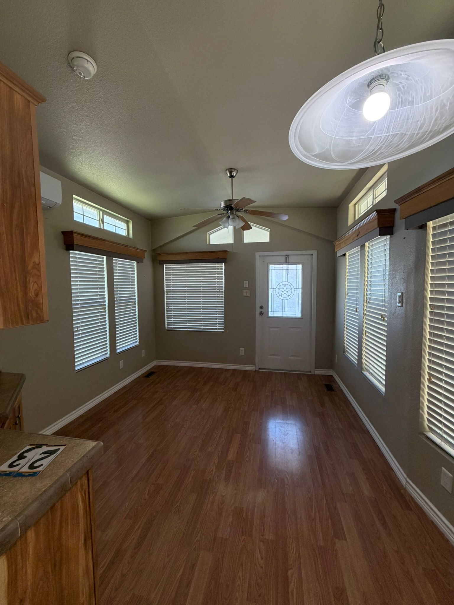 Empty living-dining room with wood floors, beige walls, ceiling fan, and large windows with blinds