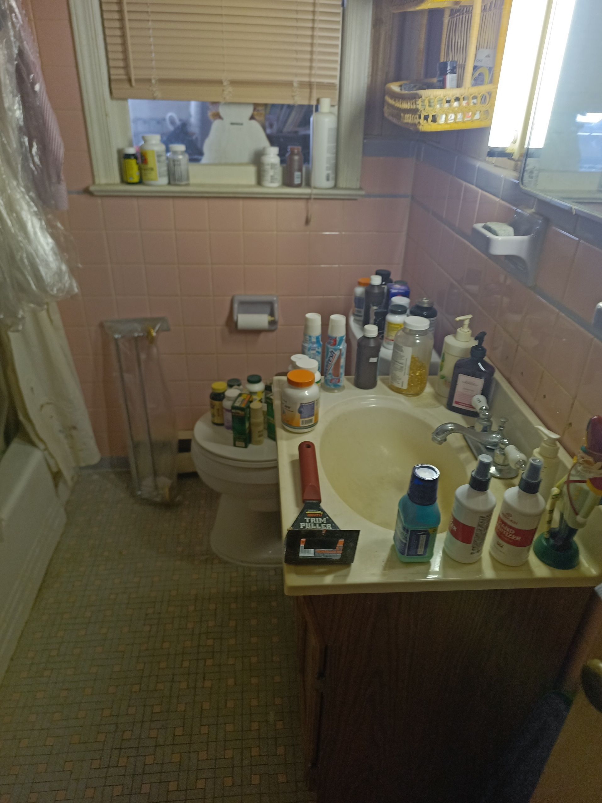 Small bathroom with a toilet, sink crowded with toiletries, and a window with blinds.