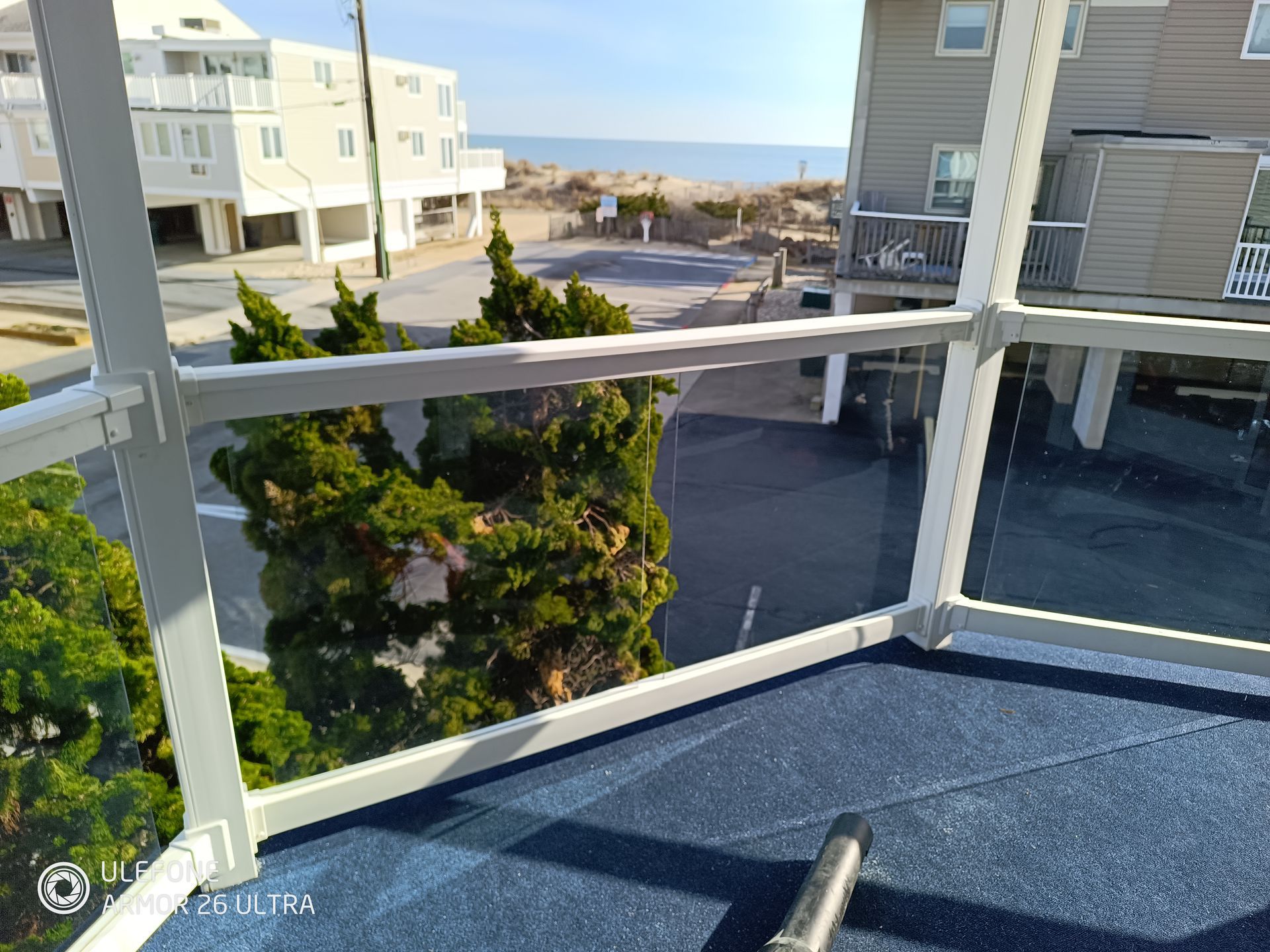 View from a balcony overlooking a beach, road, and houses. White railing surrounds a blue surface.