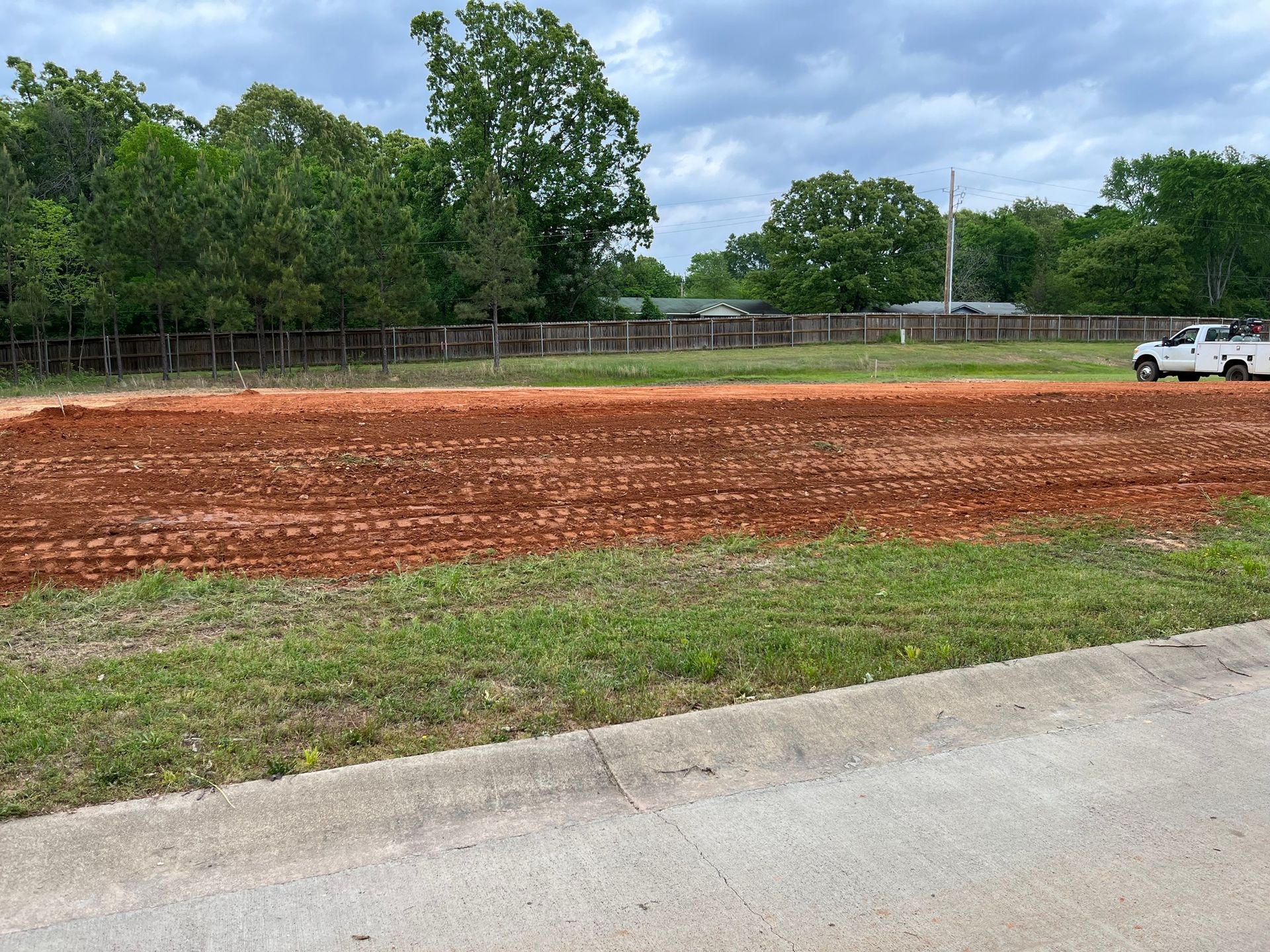 Brown dirt patch graded, roadside view. White truck parked, fence and trees in background. Cloudy sky.