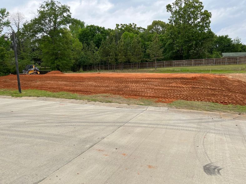 A leveled, reddish-brown dirt lot, prepared for construction, with a background of green trees and cloudy sky.