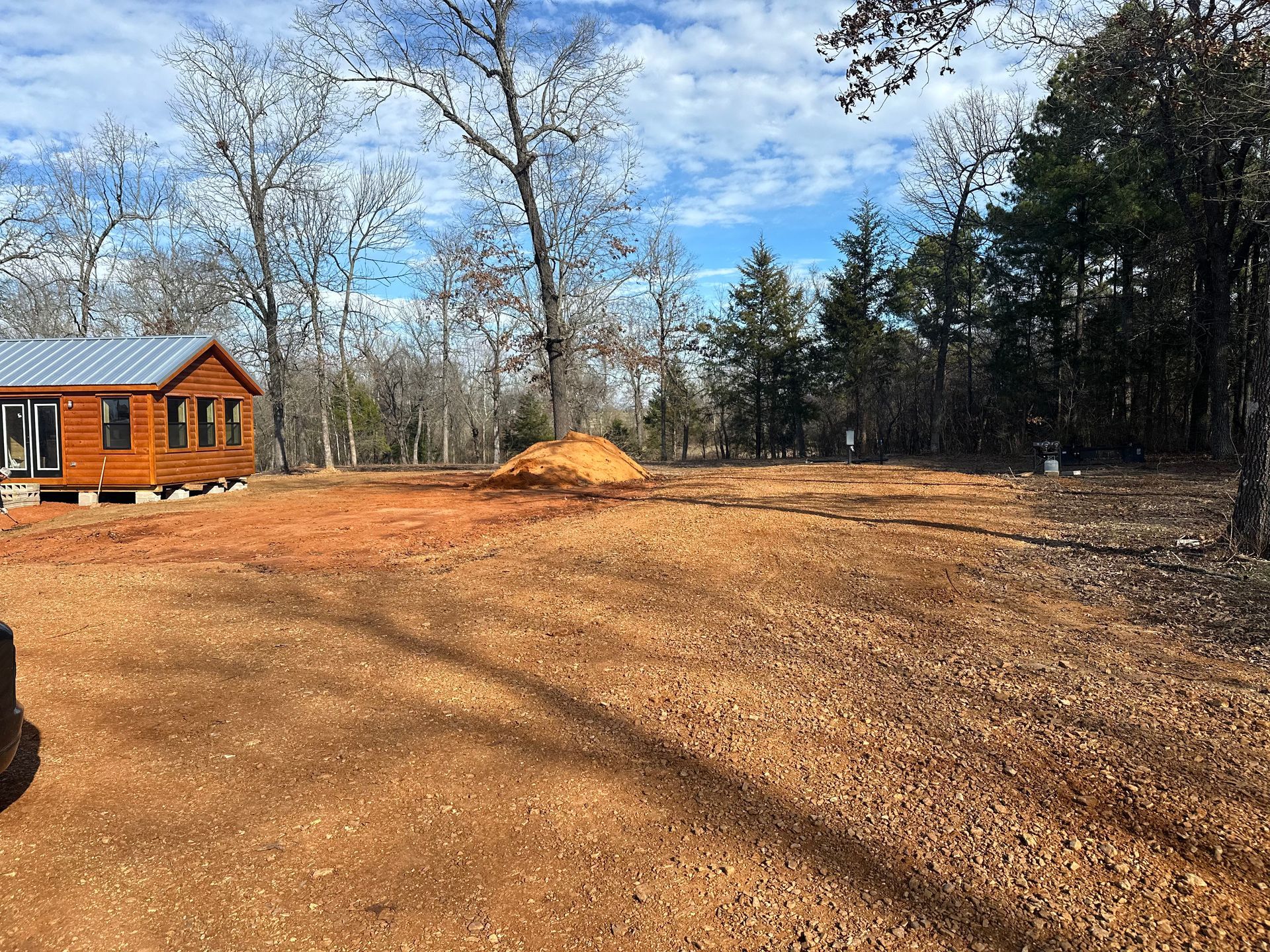 A cleared lot with reddish-brown dirt, small dirt mound, and a cabin in the background. Trees surround.