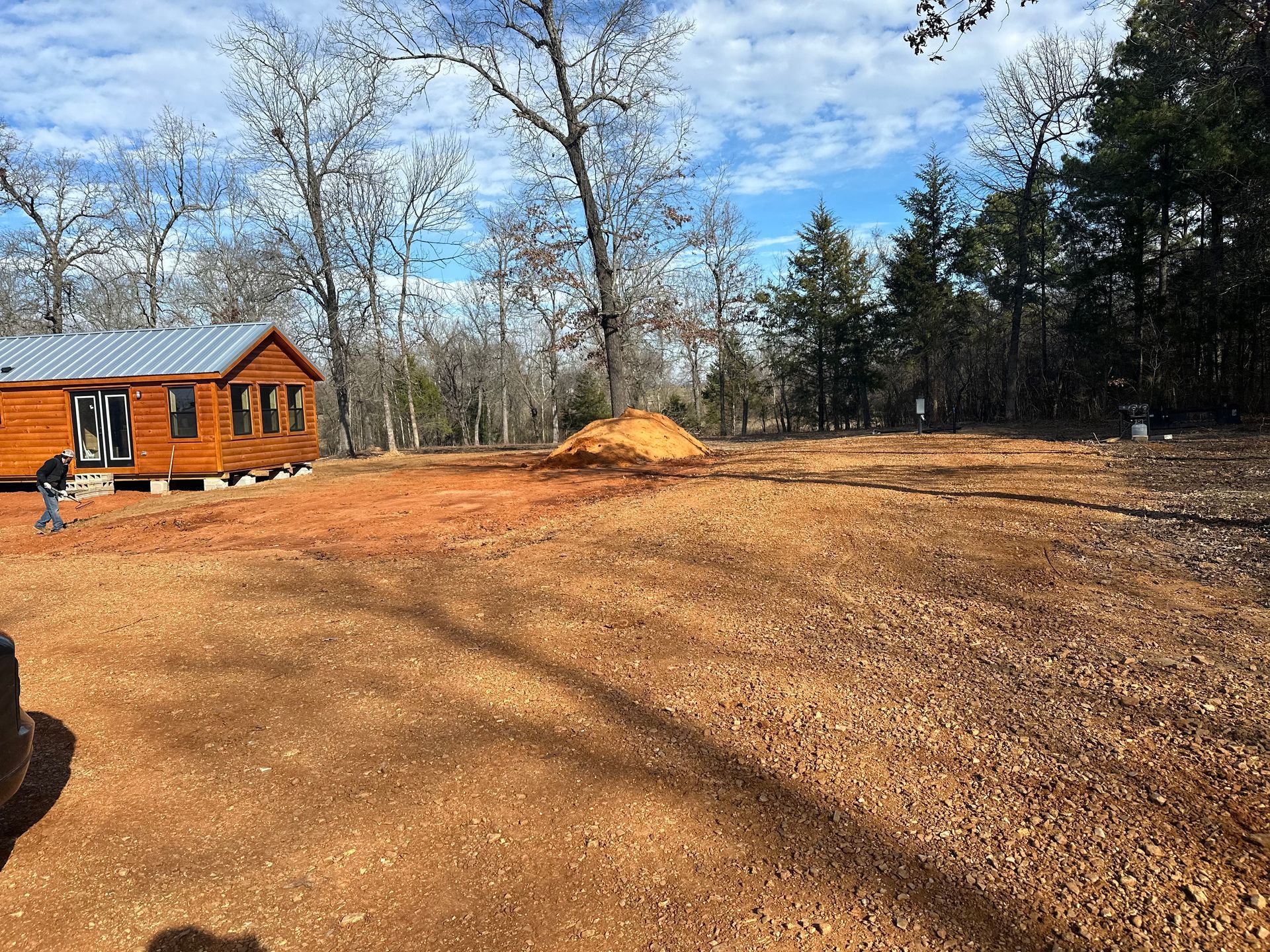 Cleared land with a cabin, dirt pile, and trees under a blue sky.