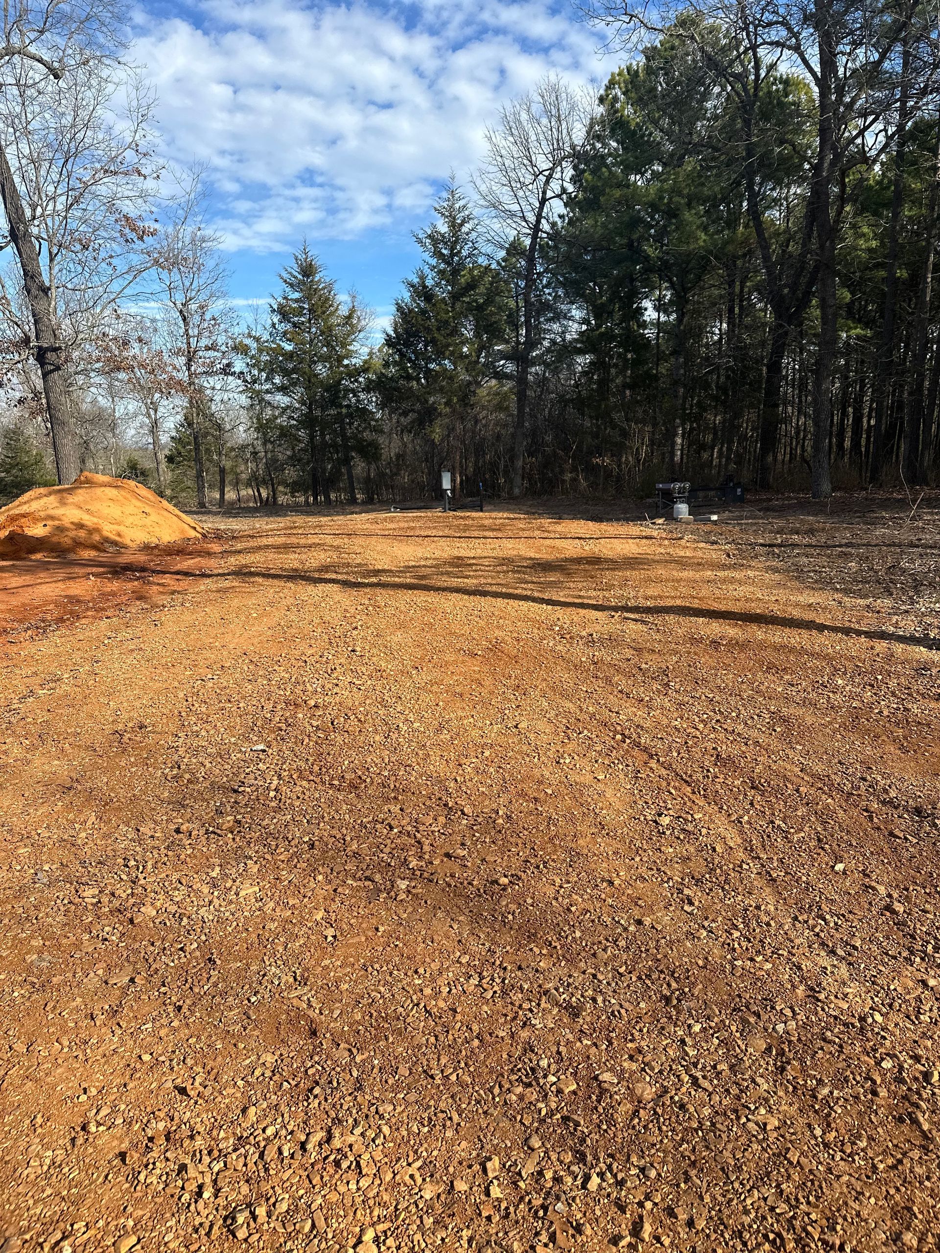 Gravel lot with a small dirt pile and trees under a partly cloudy blue sky.