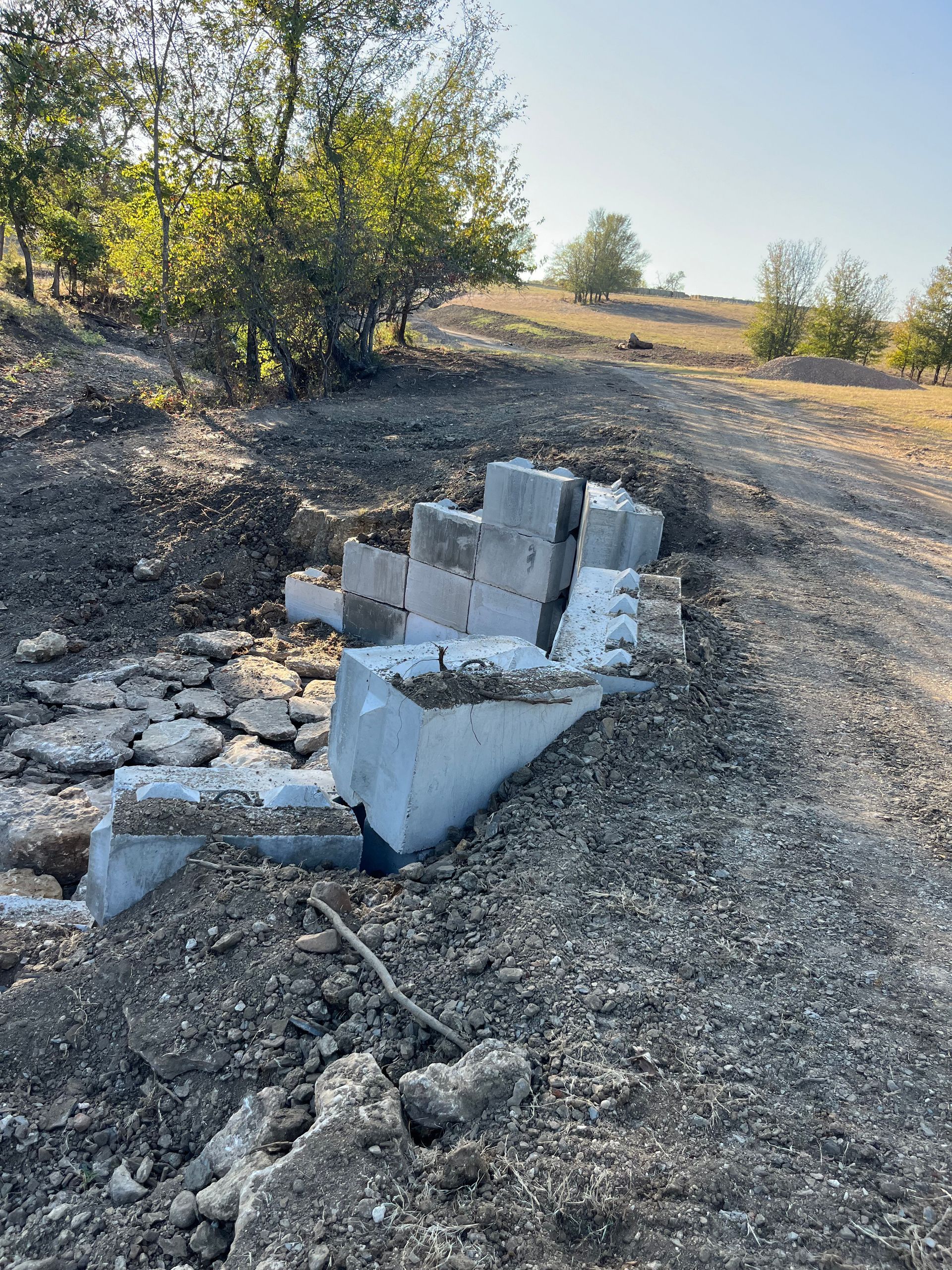 Damaged concrete blocks piled in dirt on a hillside, next to trees, under a bright sky.