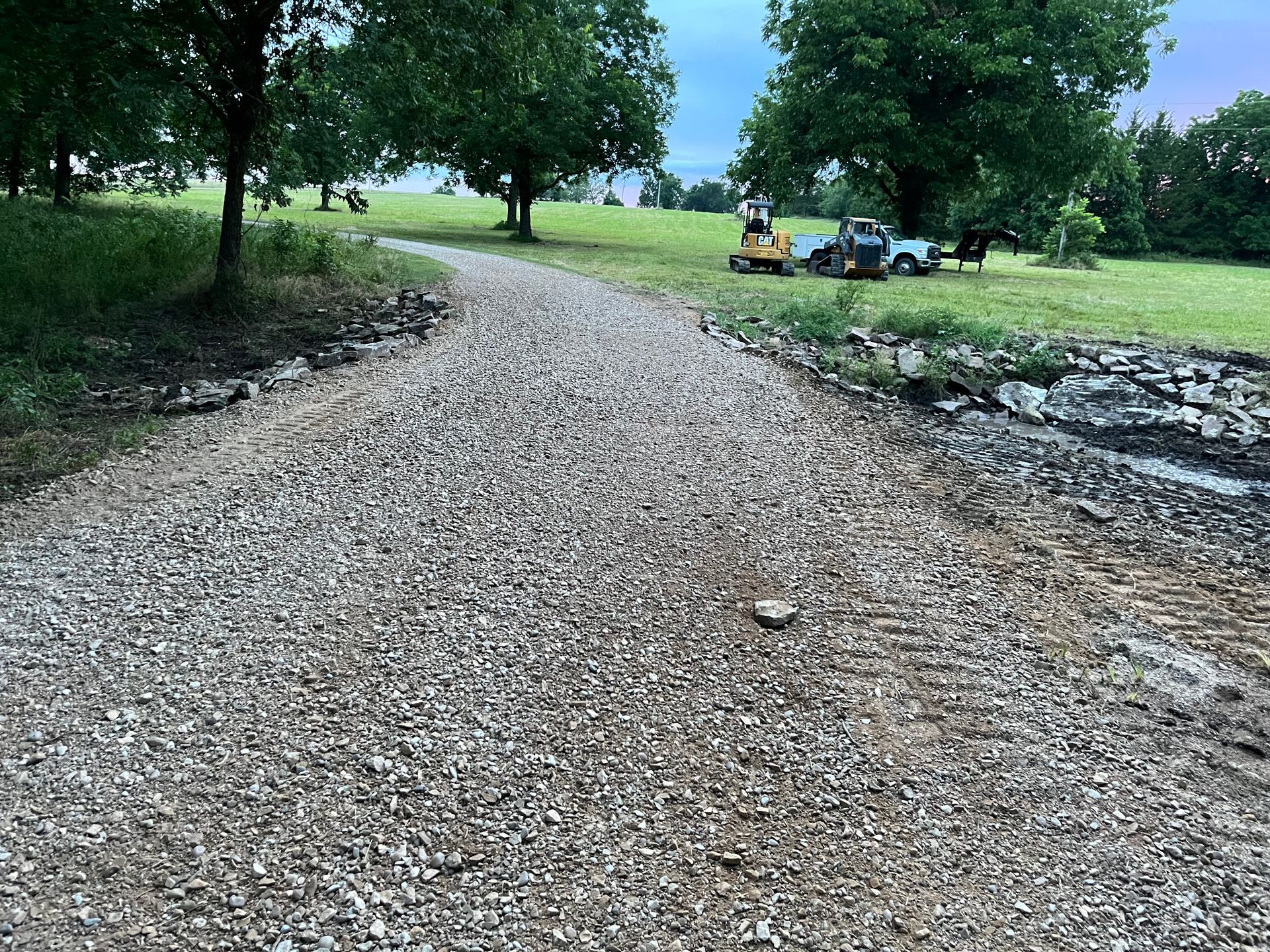 Gravel driveway leading through a green field, trees in background, construction equipment nearby.