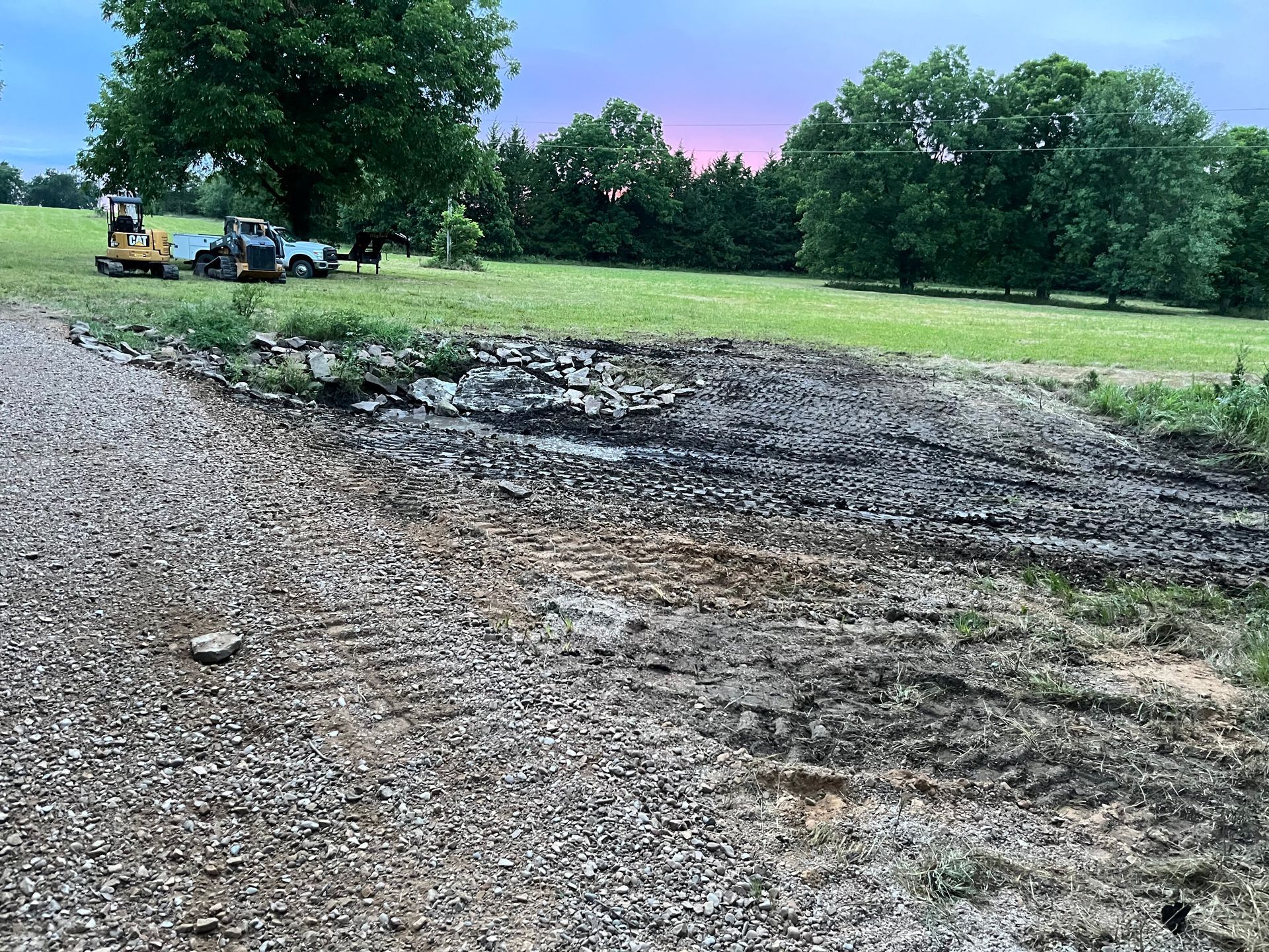 Gravel driveway and dirt path with a machine and truck in the background, trees under a dusky sky.
