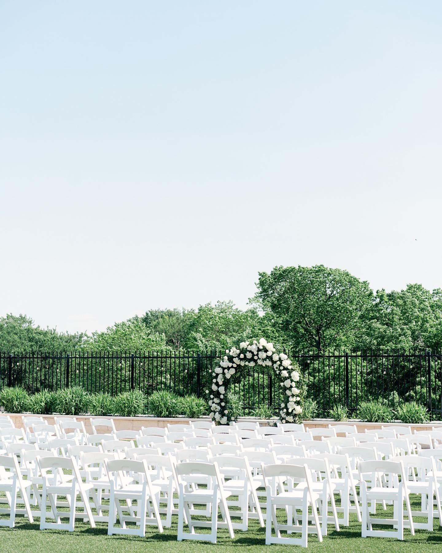 A row of white chairs are lined up in a field with a floral arch in the background.