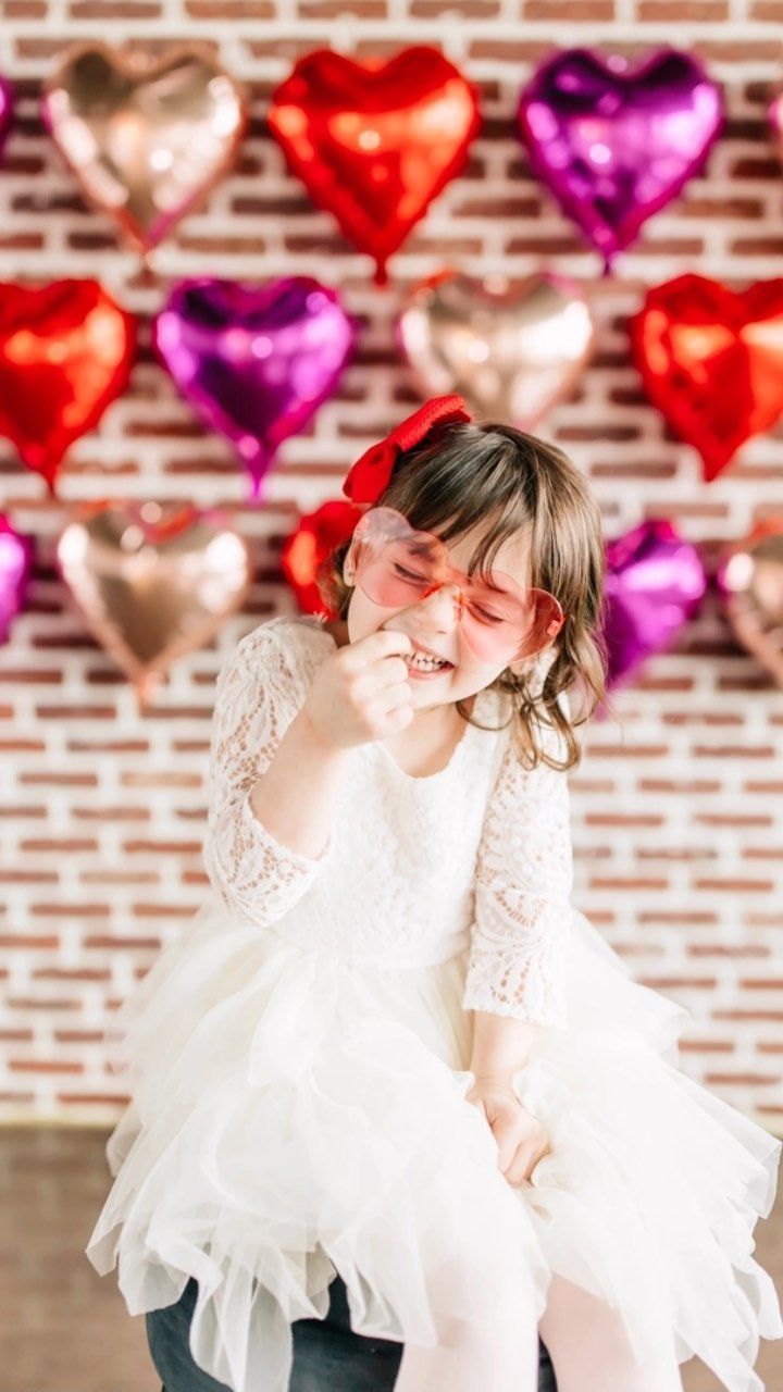 A little girl in a white dress is sitting in front of a wall of heart shaped balloons.