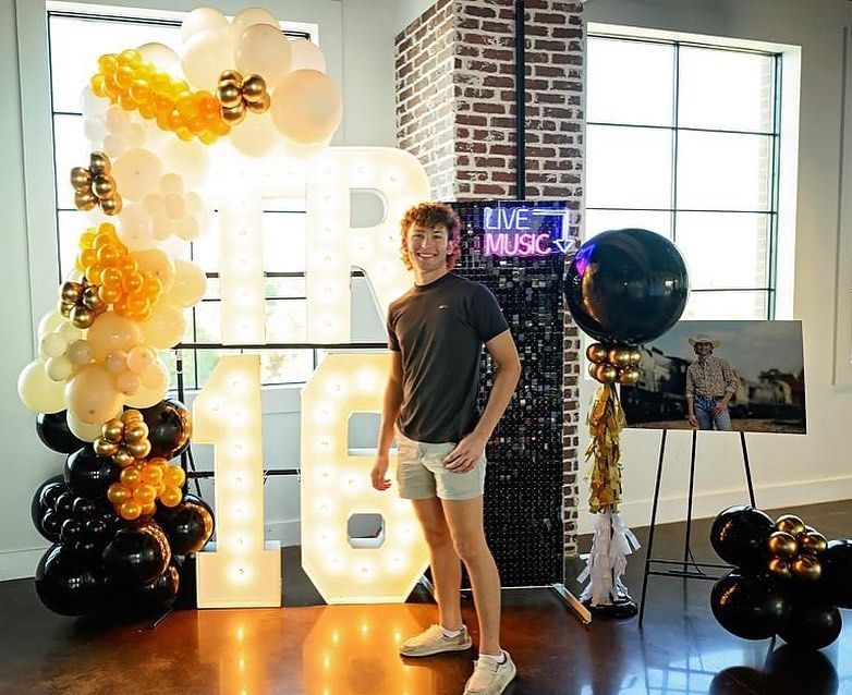 A young man is standing in front of a large r surrounded by balloons.