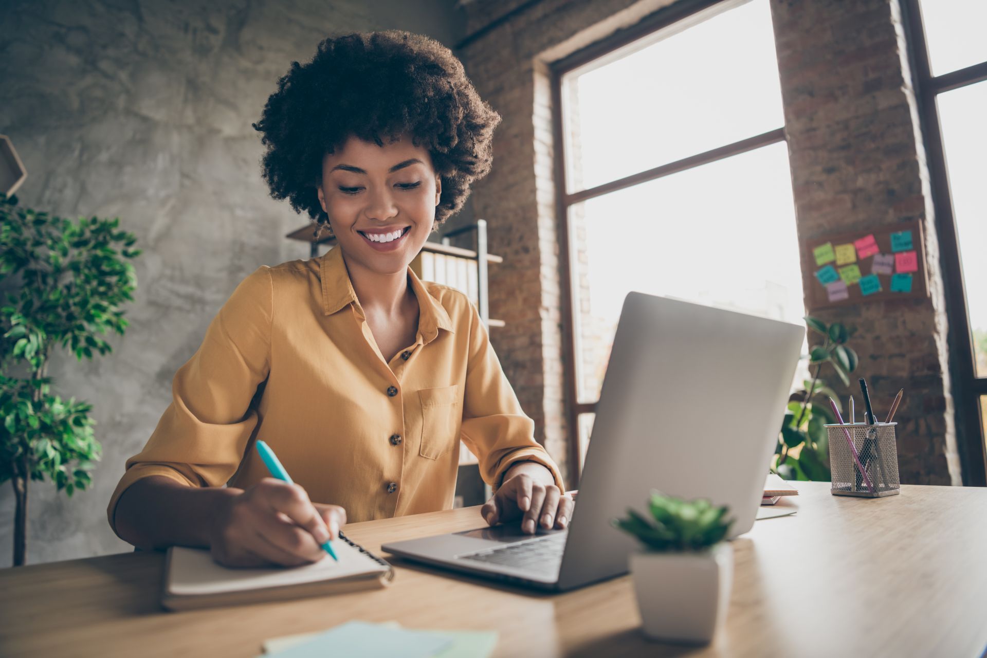 Woman in yellow shirt smiles, writing in notebook while using laptop at a desk. Sunlight streams in from a window.