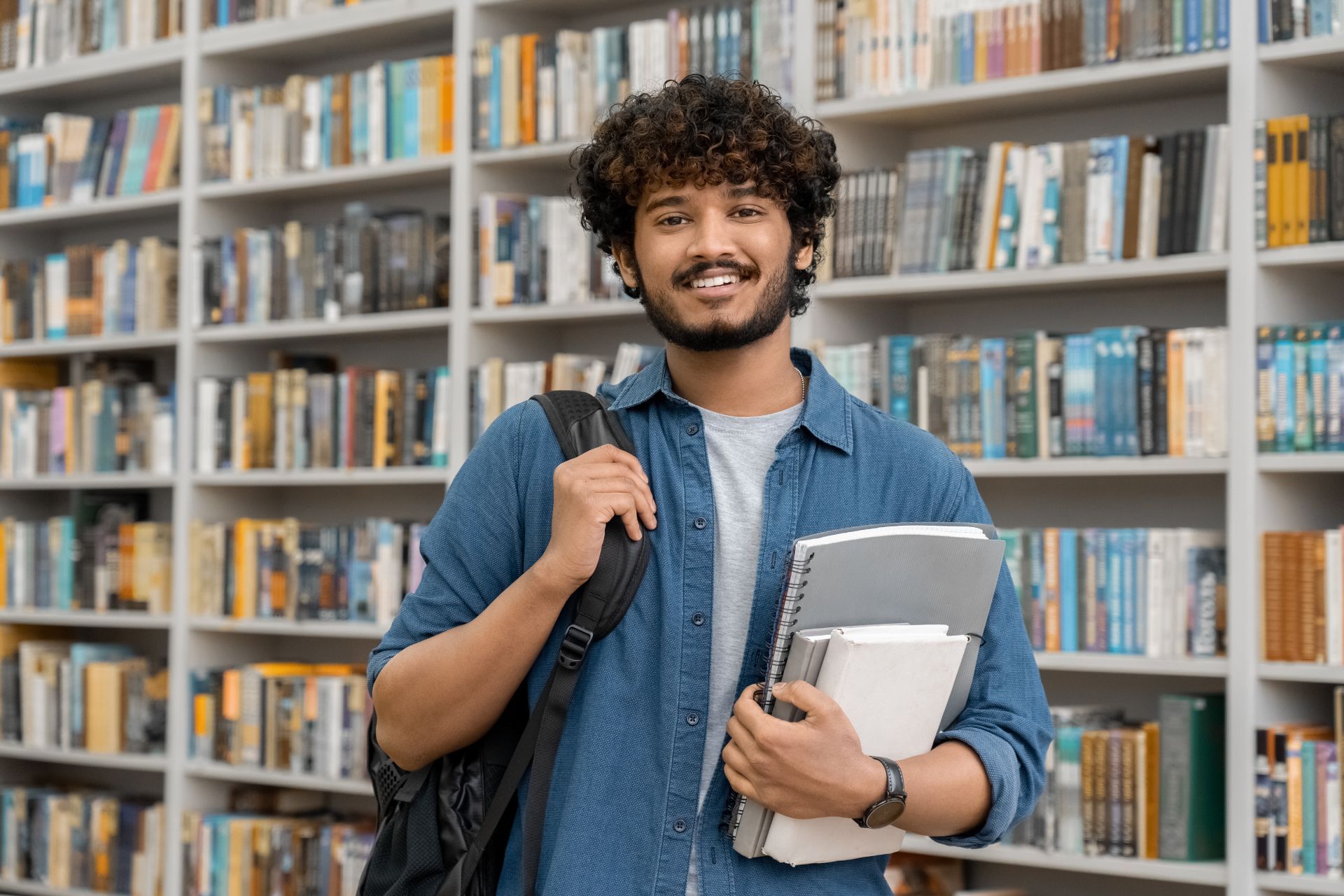 Man in denim shirt with backpack and books smiles in a library.