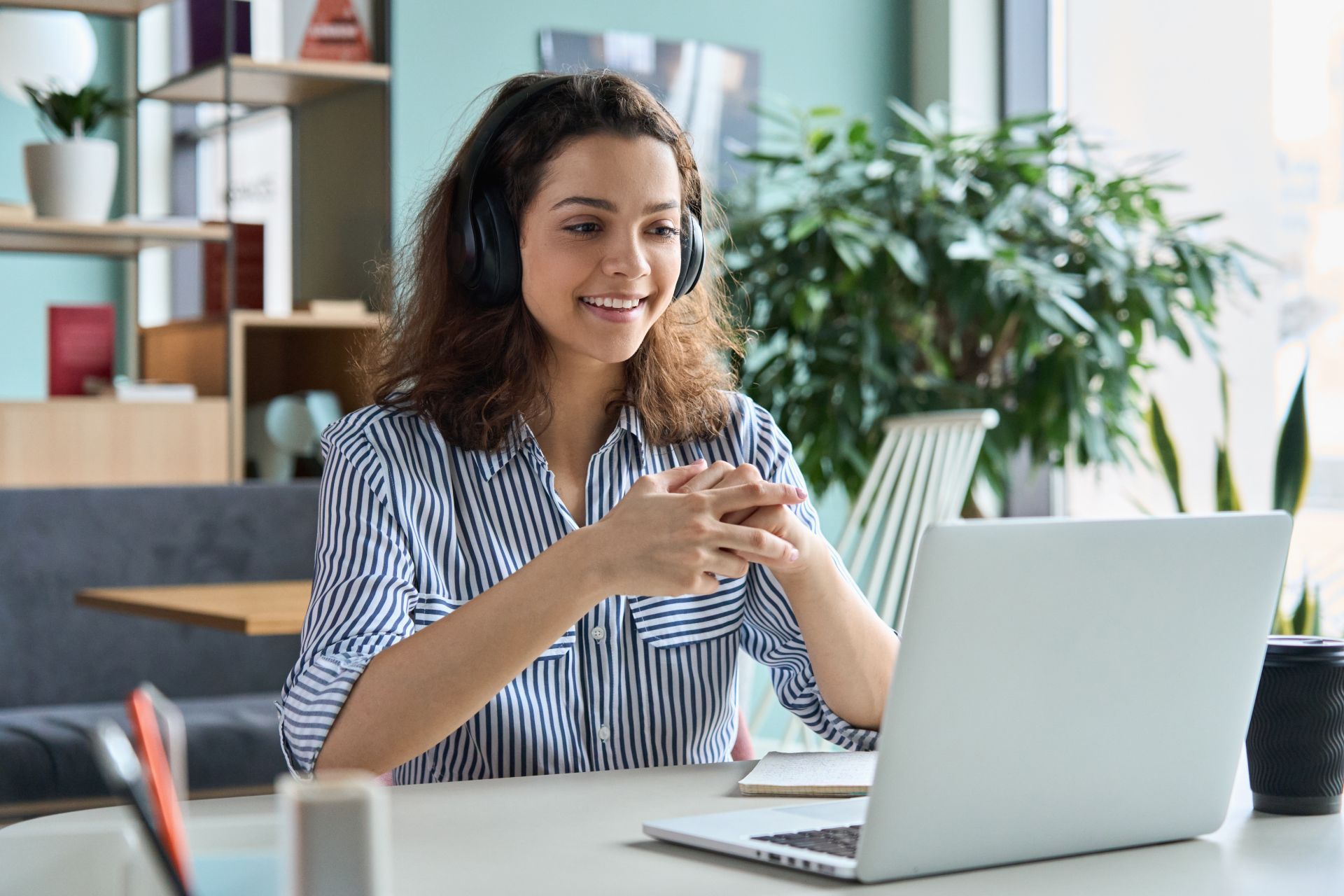 Woman wearing headphones, smiling at a laptop, hands clasped, in a cafe setting.