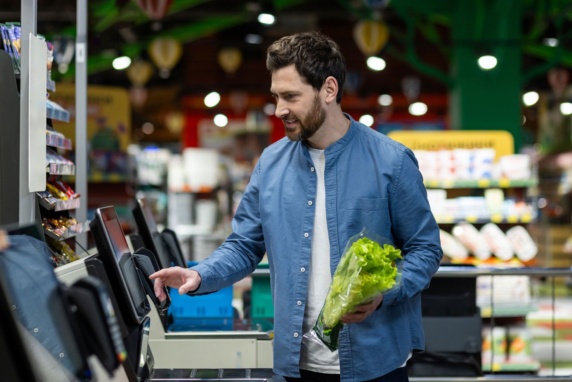 Man scanning lettuce at a self-checkout in a grocery store. He is smiling, with a blue shirt.