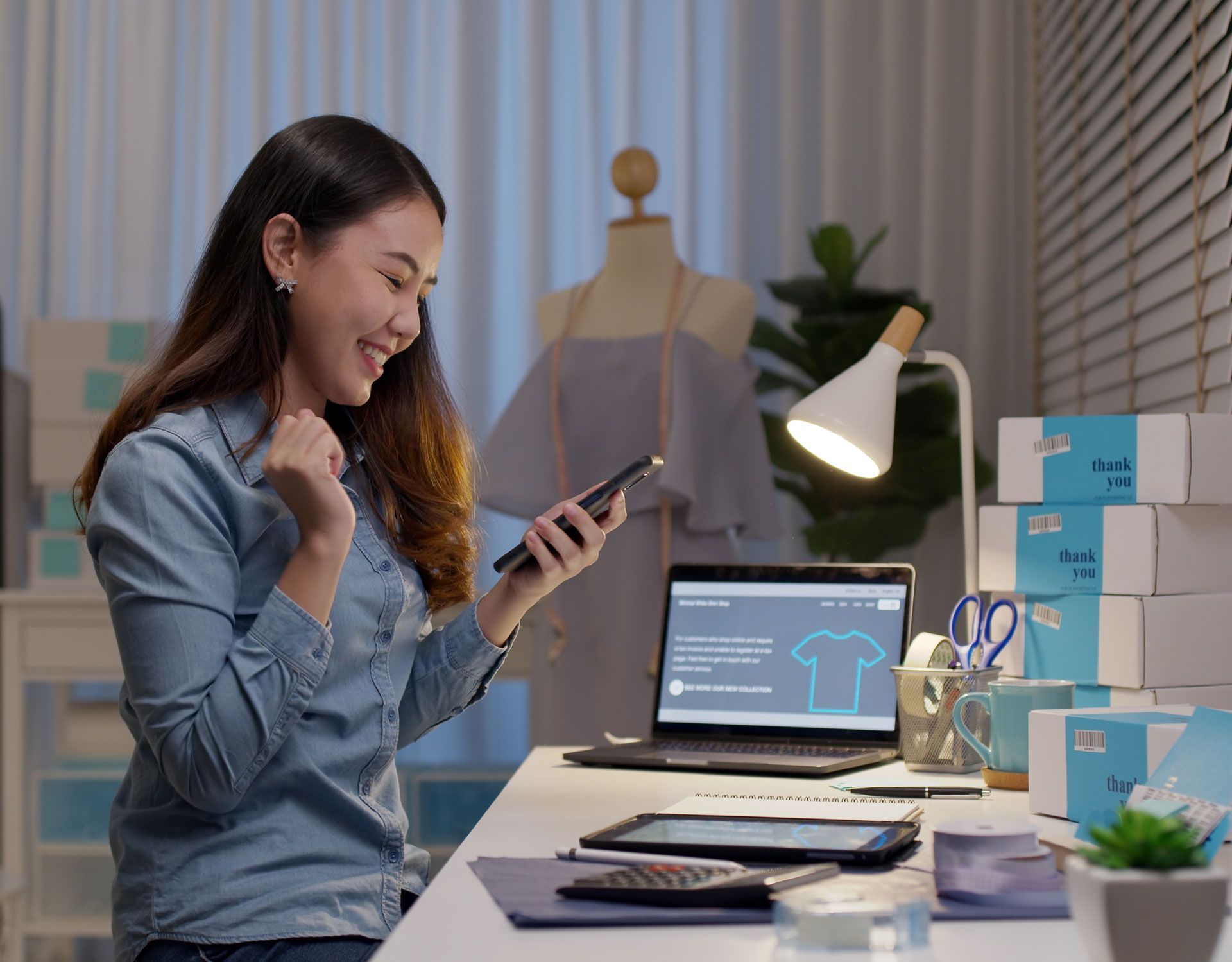 Woman smiles, looking at phone. She works at a desk, surrounded by boxes and a laptop.
