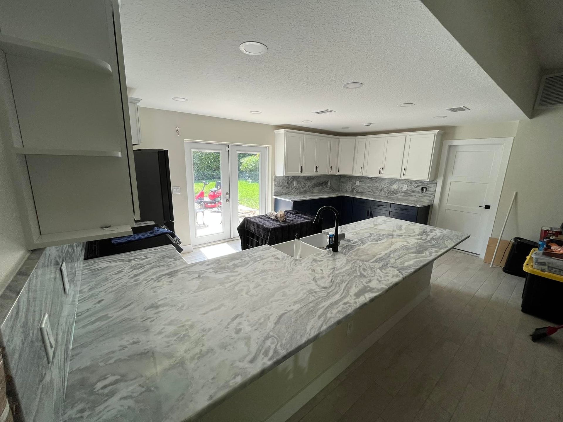 A modern kitchen with white upper cabinets, dark base cabinets, gray-and-white stone countertops, and glass patio doors.