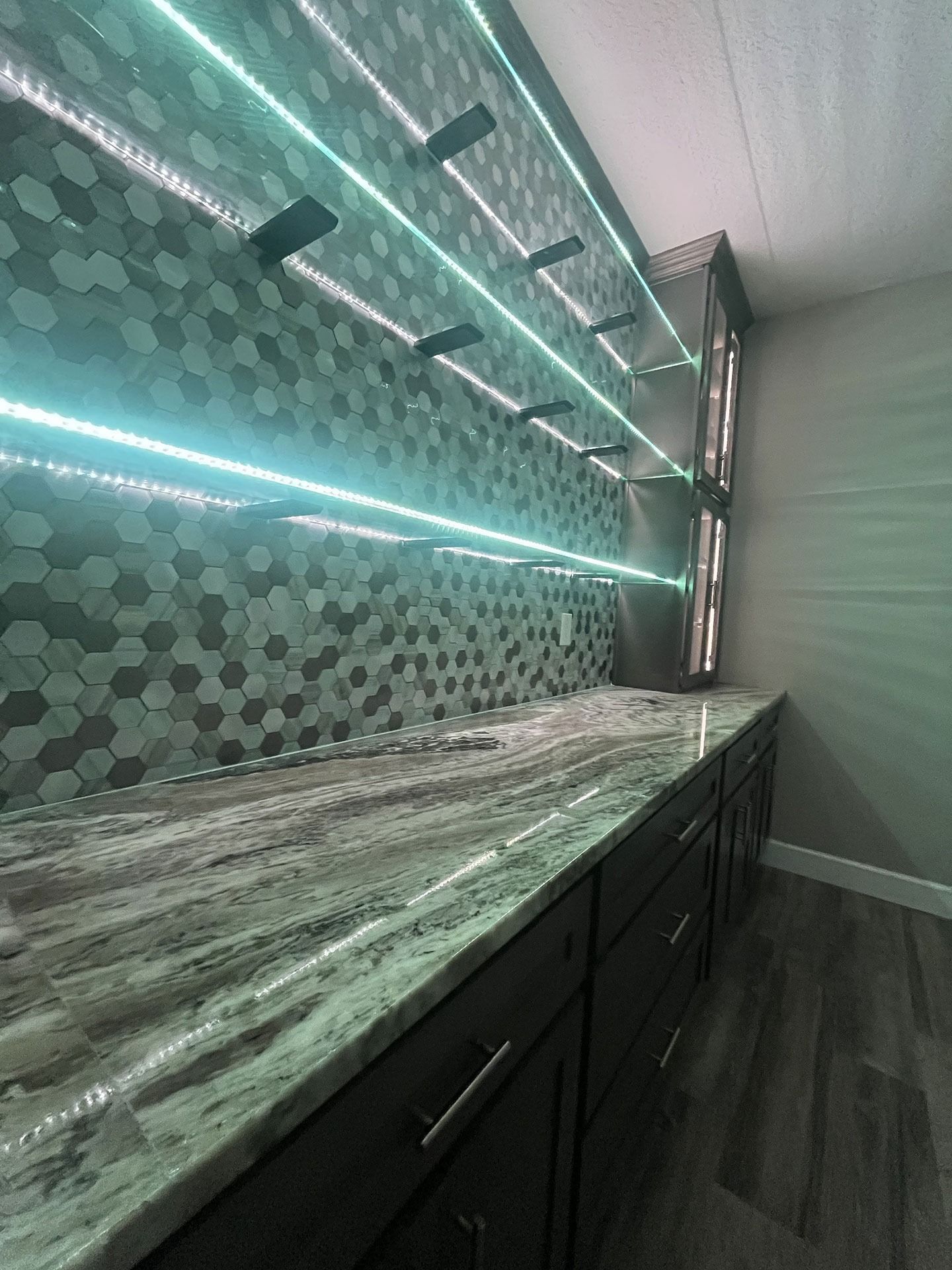 A kitchen counter with a stone countertop, a hexagonal tile backsplash, and three horizontal floating shelves with LEDs.