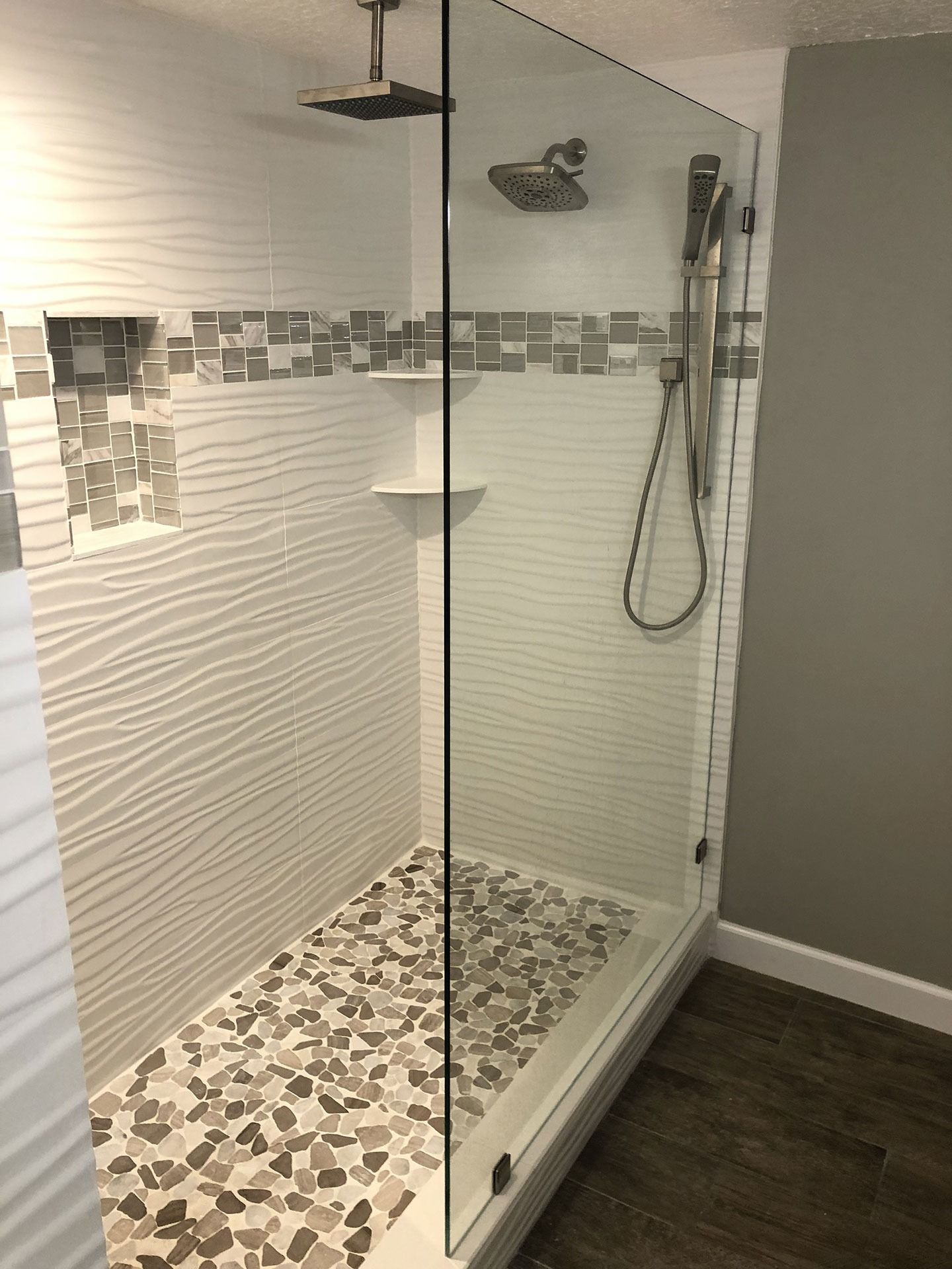 A modern walk-in shower with textured white tile walls, a pebble floor, a glass partition, and two shower heads.