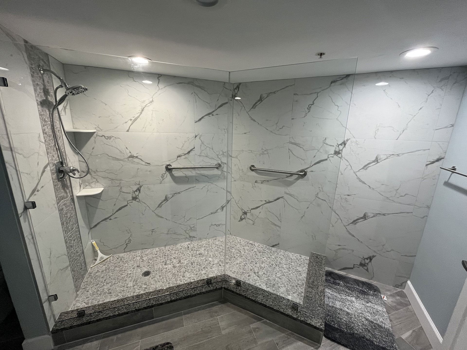 A wheelchair-accessible shower with white marble-patterned walls, stone-tiled floor, grab bars, and a corner bench.