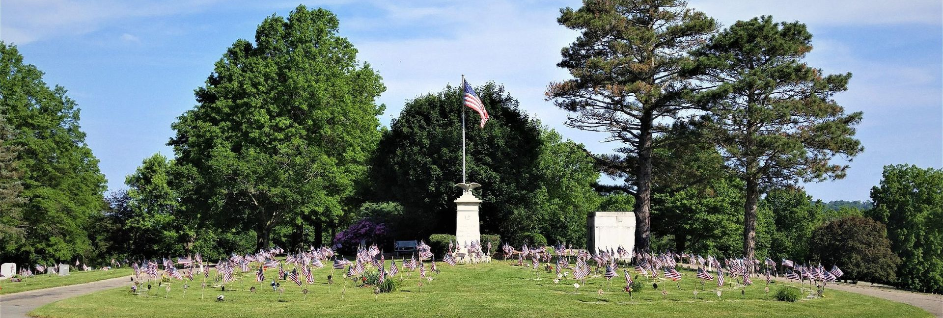 Grassy memorial garden with a white statue, flowers, and tall green trees under a bright sky