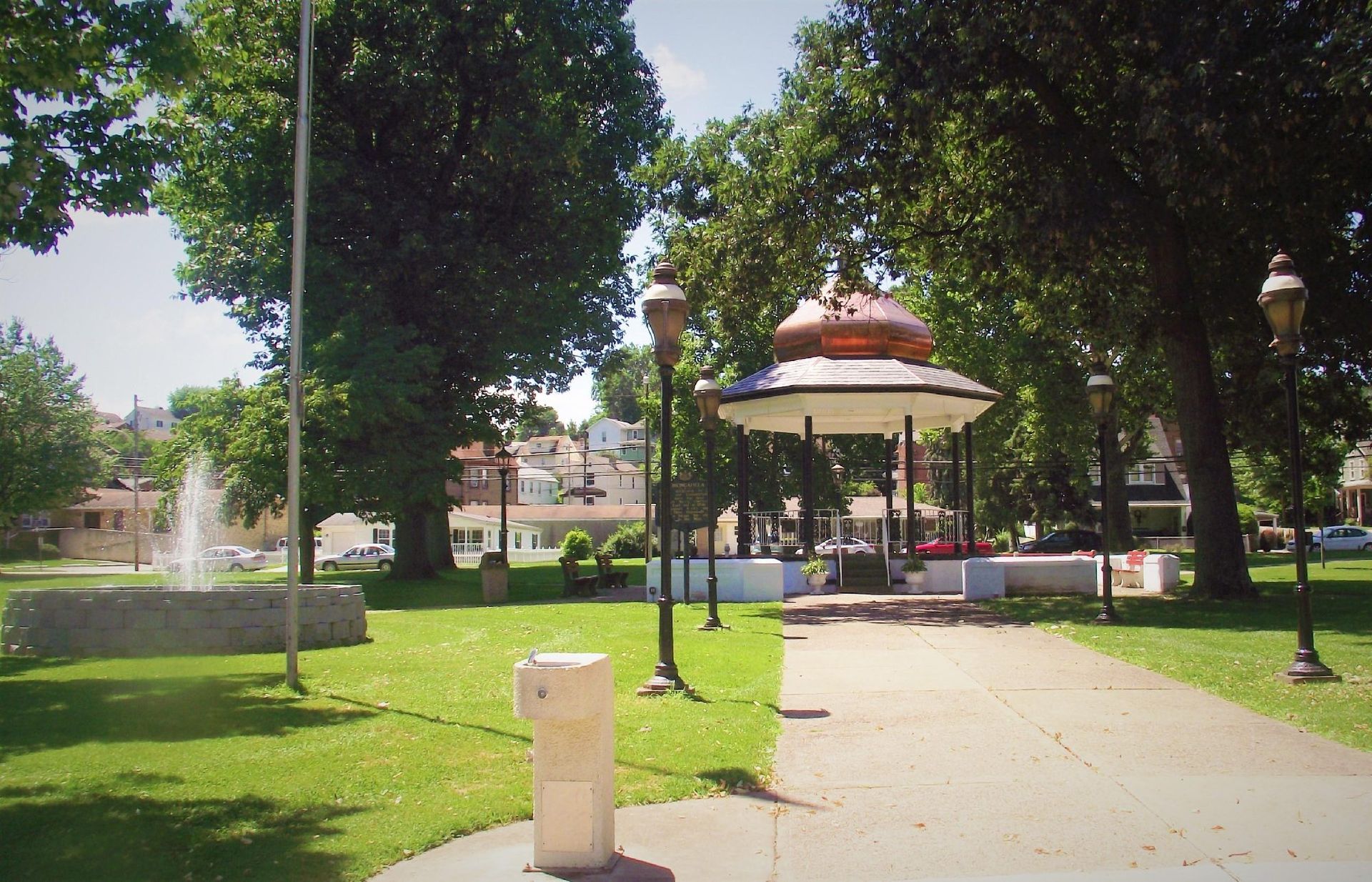 Park with trees, gazebo, fountain, and a paved path on a sunny day.