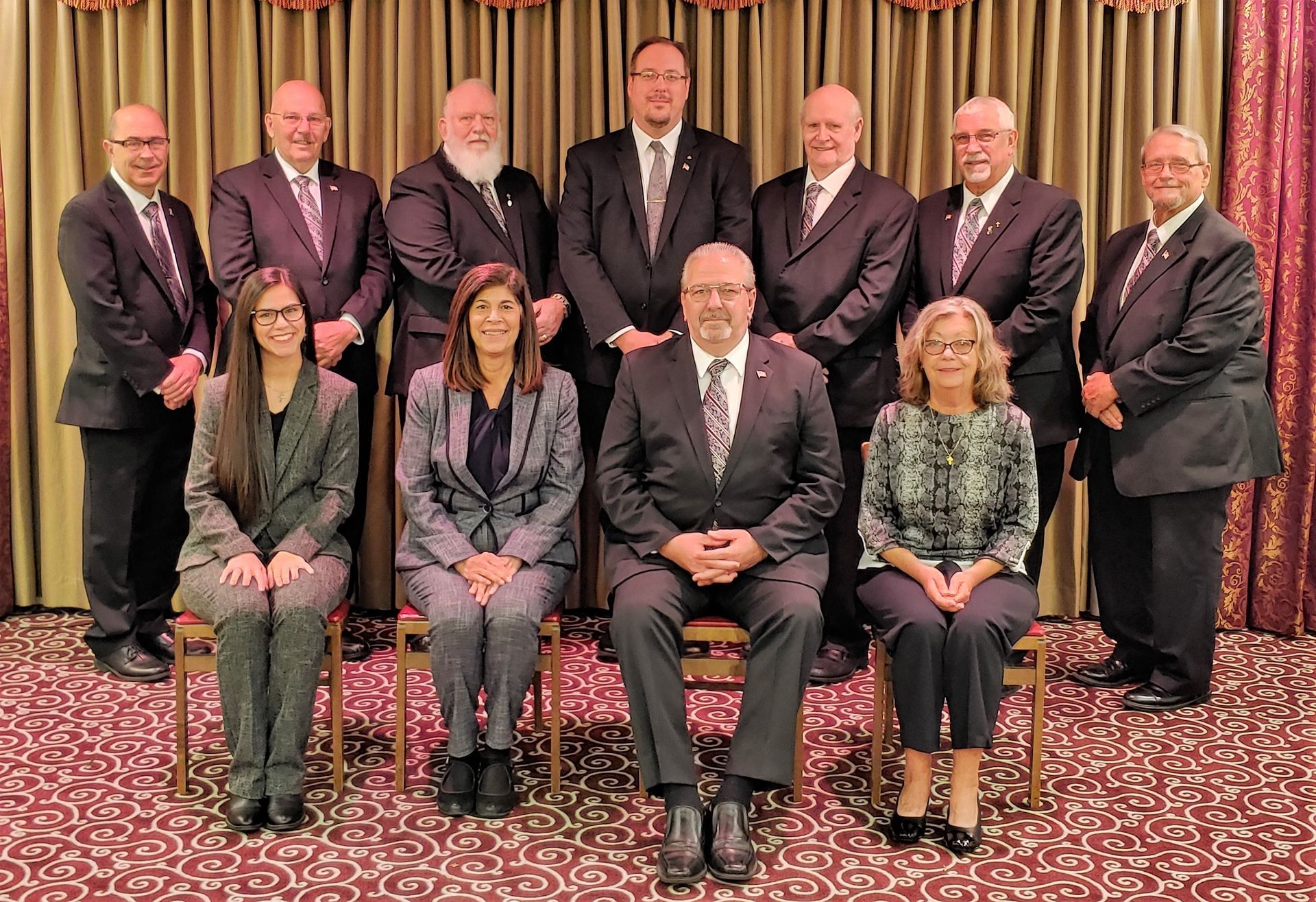 Group of people posing for a formal team portrait in suits and dresses on a carpeted stage.
