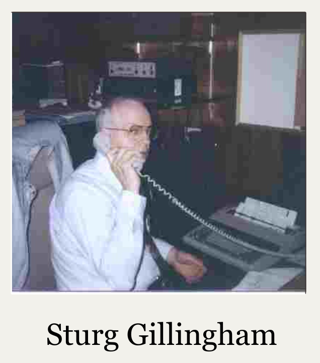 Man seated at a desk holding a corded phone in a dim office, with the name