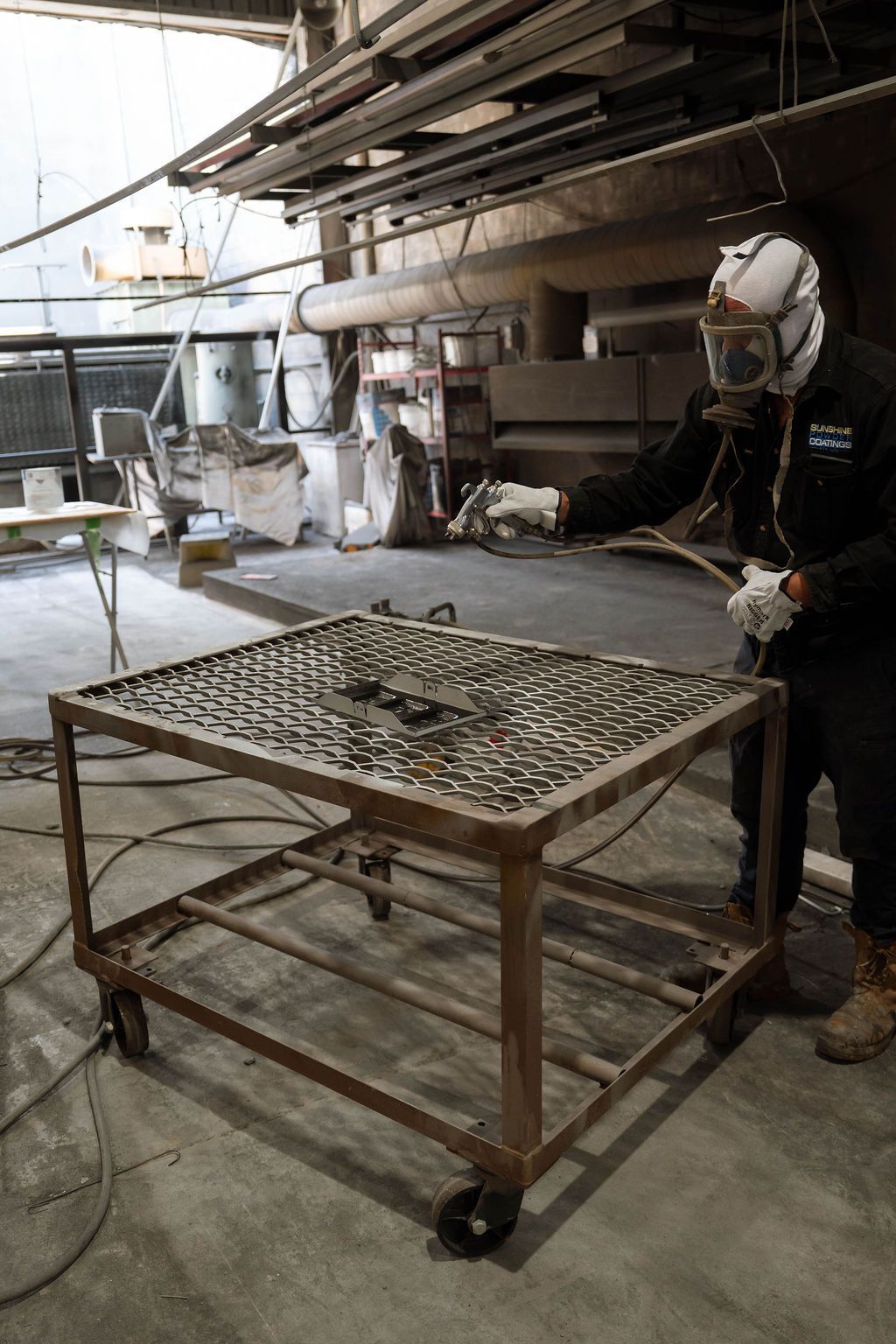 A worker in protective gear and a respirator uses a spray gun on metal parts arranged on a rolling industrial cart.
