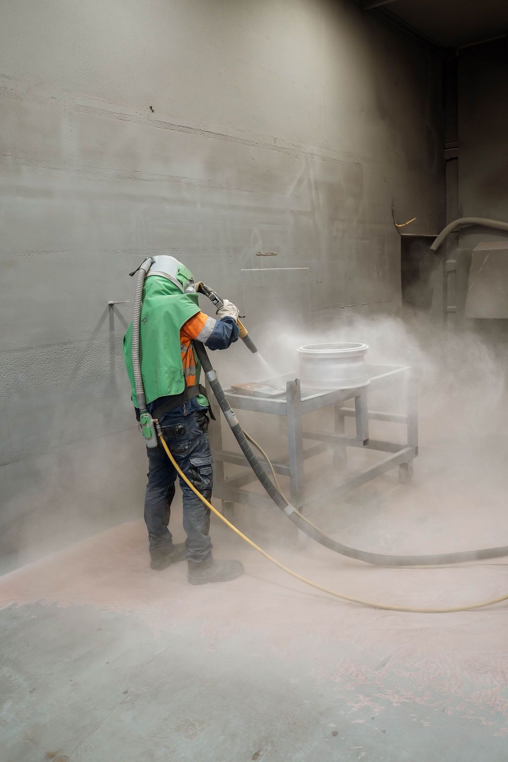 A worker in protective gear uses a blast nozzle to sandblast a metal object in an industrial room filled with dust.