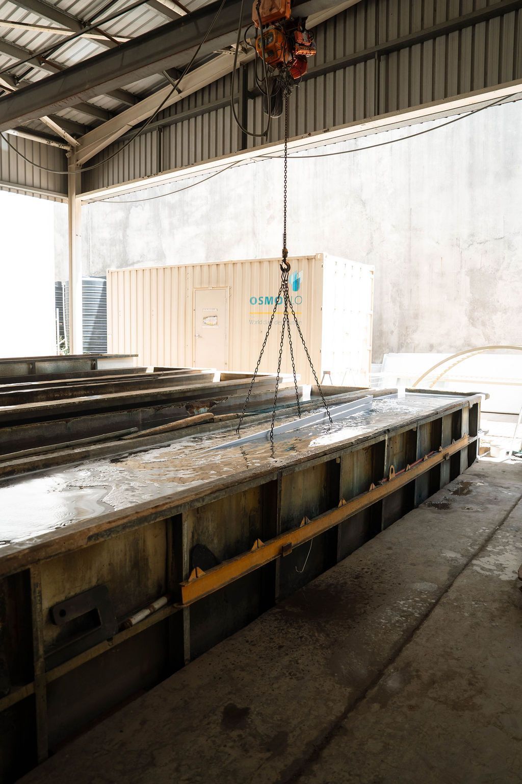 An overhead crane lowers a metal frame into a long, industrial water-filled trough inside a warehouse.