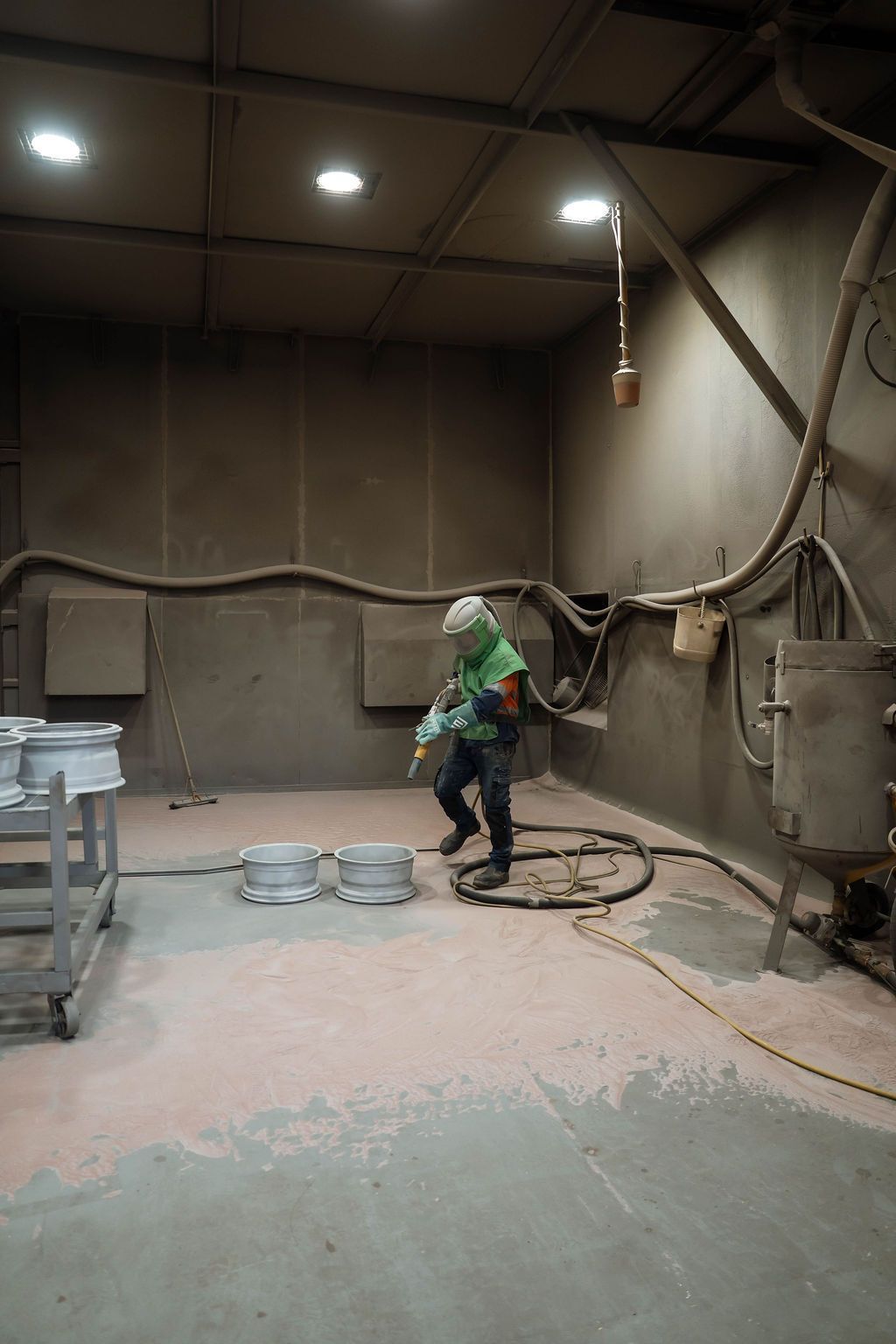 A worker in a protective suit sandblasts metal wheels inside a dusty, industrial workshop.