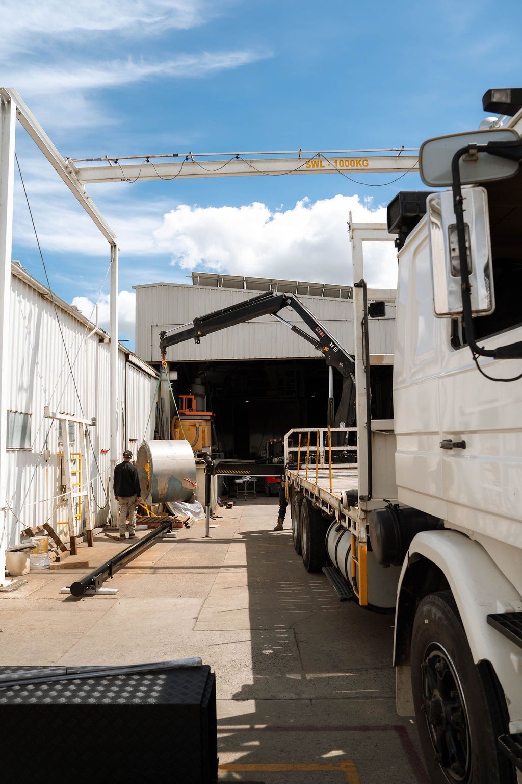 A truck with a mounted crane sits in an industrial yard near a large metal coil under a bright, cloudy sky.