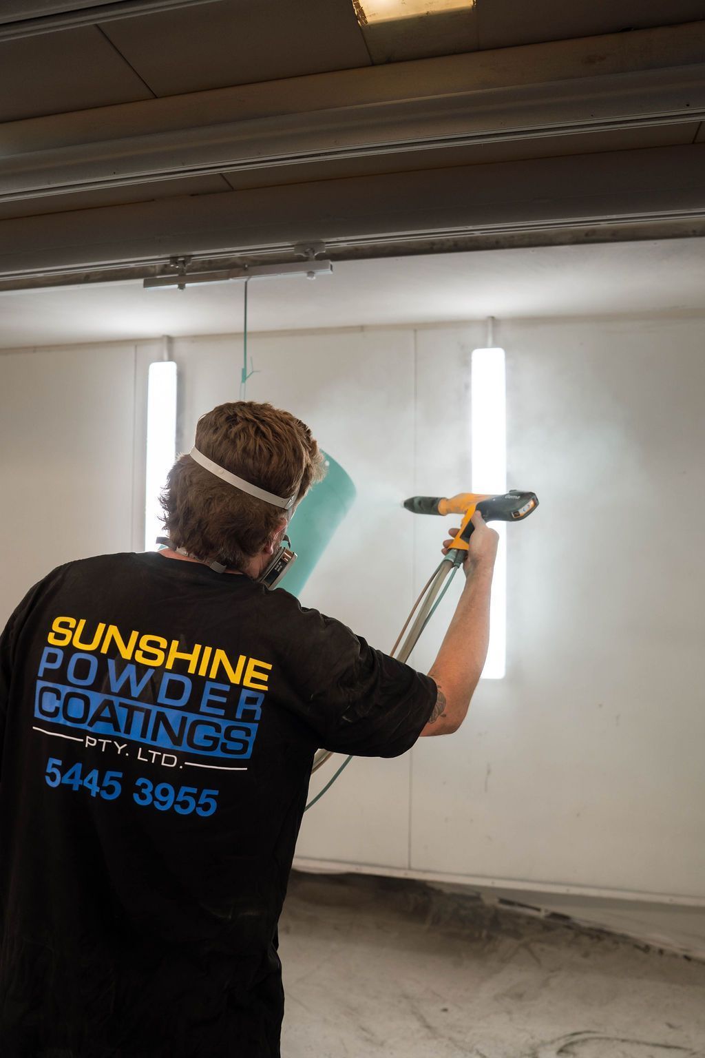 A person wearing a Sunshine Powder Coatings shirt uses a spray gun to apply powder coating to a part in a bright booth.