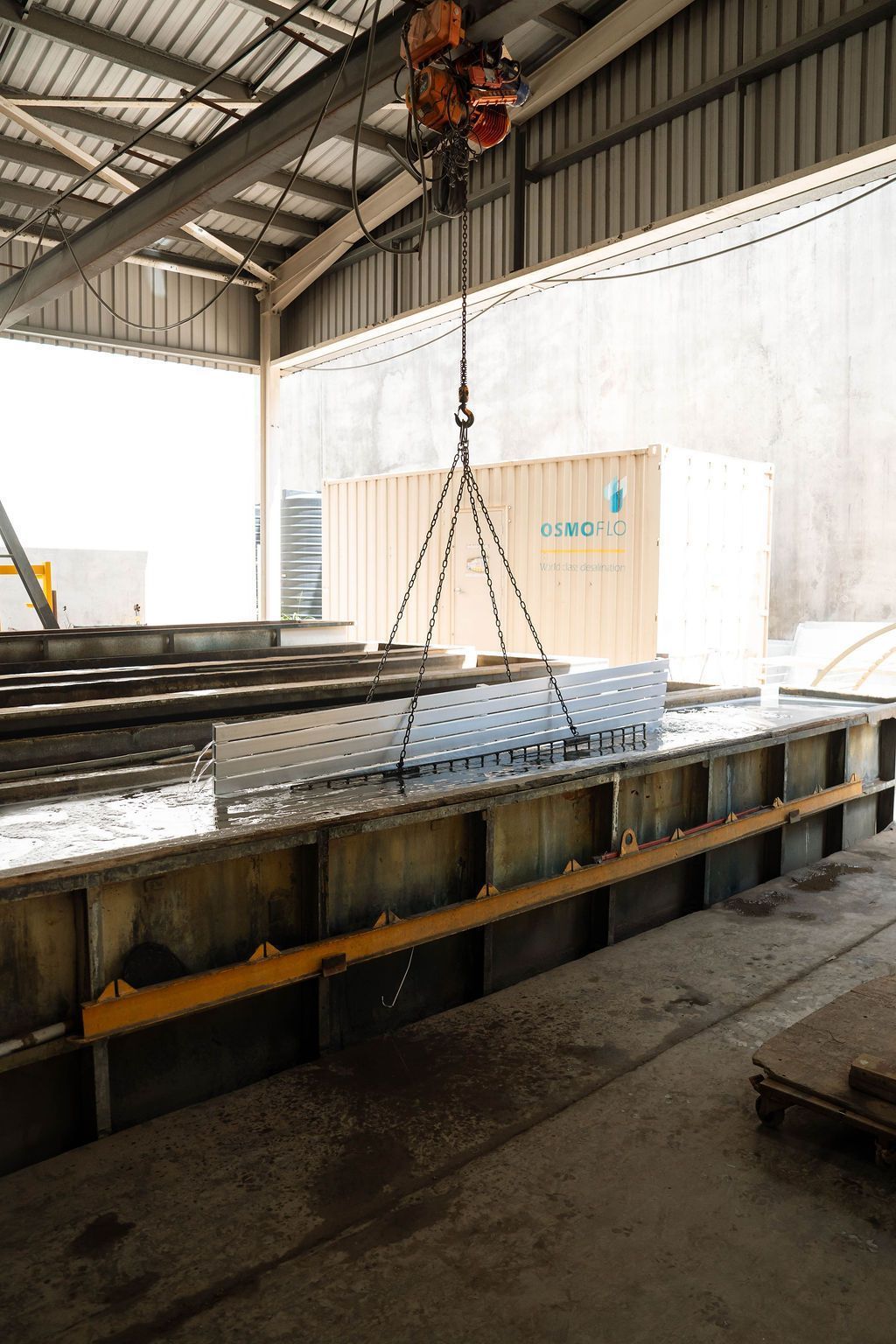 A crane hoist lifts a metal perforated panel over a long rectangular industrial tank inside a warehouse.